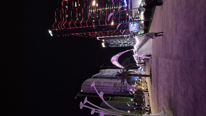 Night view of city buildings and waterfront area illuminated in colorful lights. A tall, modern building with red vertical lights dominates the upper left. A curved, illuminated walkway spans across the lower area, surrounded by palm trees and lower buildings. A concrete promenade extends towards the right, where a couple of people are visible. The overall impression is of a vibrant, modern urban landscape at night.
