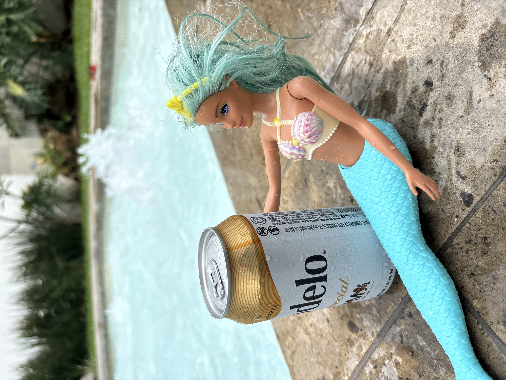The image depicts a mermaid doll with light blue hair and a seashell bikini top, lying next to a can of Modelo Especial beer on a stone surface. The mermaid doll's tail is light blue and scaly. The can of beer has condensation on it. In the background, there is a body of water, possibly a pool, with a fountain feature. A blurred view of green foliage is visible on the left side of the image. The OCR data on the can indicates the product is brewed in Mexico by Cerveceria Modelo de México. The phrase "DE ESTE PRODUCTO ES NOCIVO PARA LA SALUD" appears in Spanish, which translates to "THIS PRODUCT IS HARMFUL TO YOUR HEALTH."