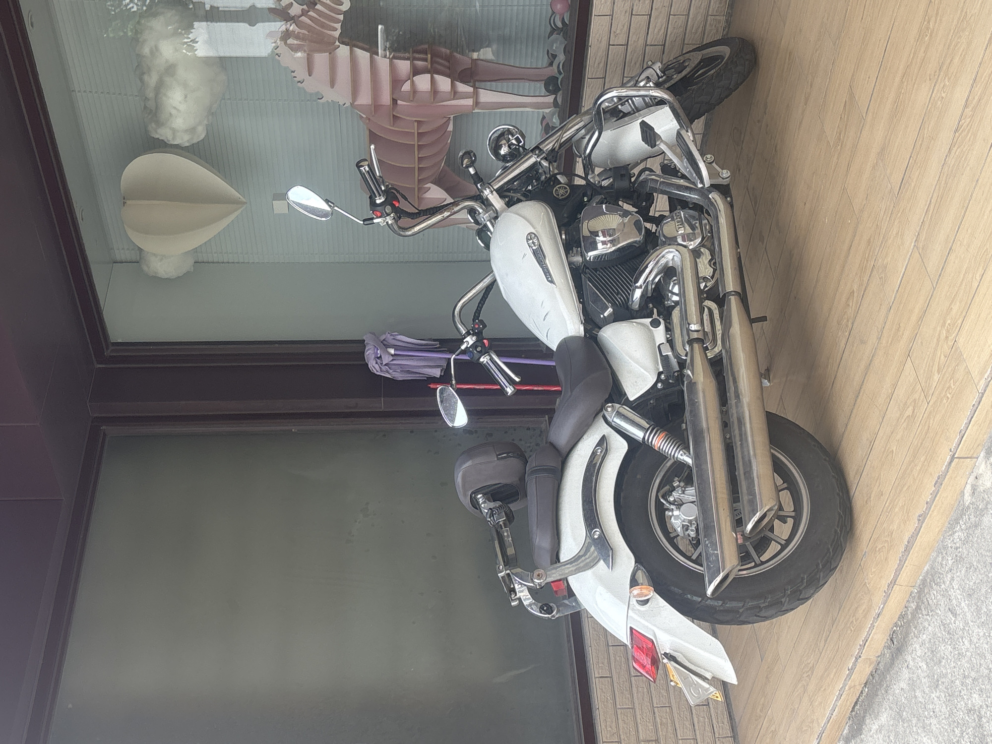 A white motorcycle is parked on a wooden tiled floor in front of a building with large windows. In the window, a large, pink, wooden cutout of a horse is visible, along with decorative paper hearts and cotton balls resembling clouds. A purple cloth is hanging on a rack to the left of the motorcycle. The scene appears to be an outdoor storefront in Nanhai District, China.