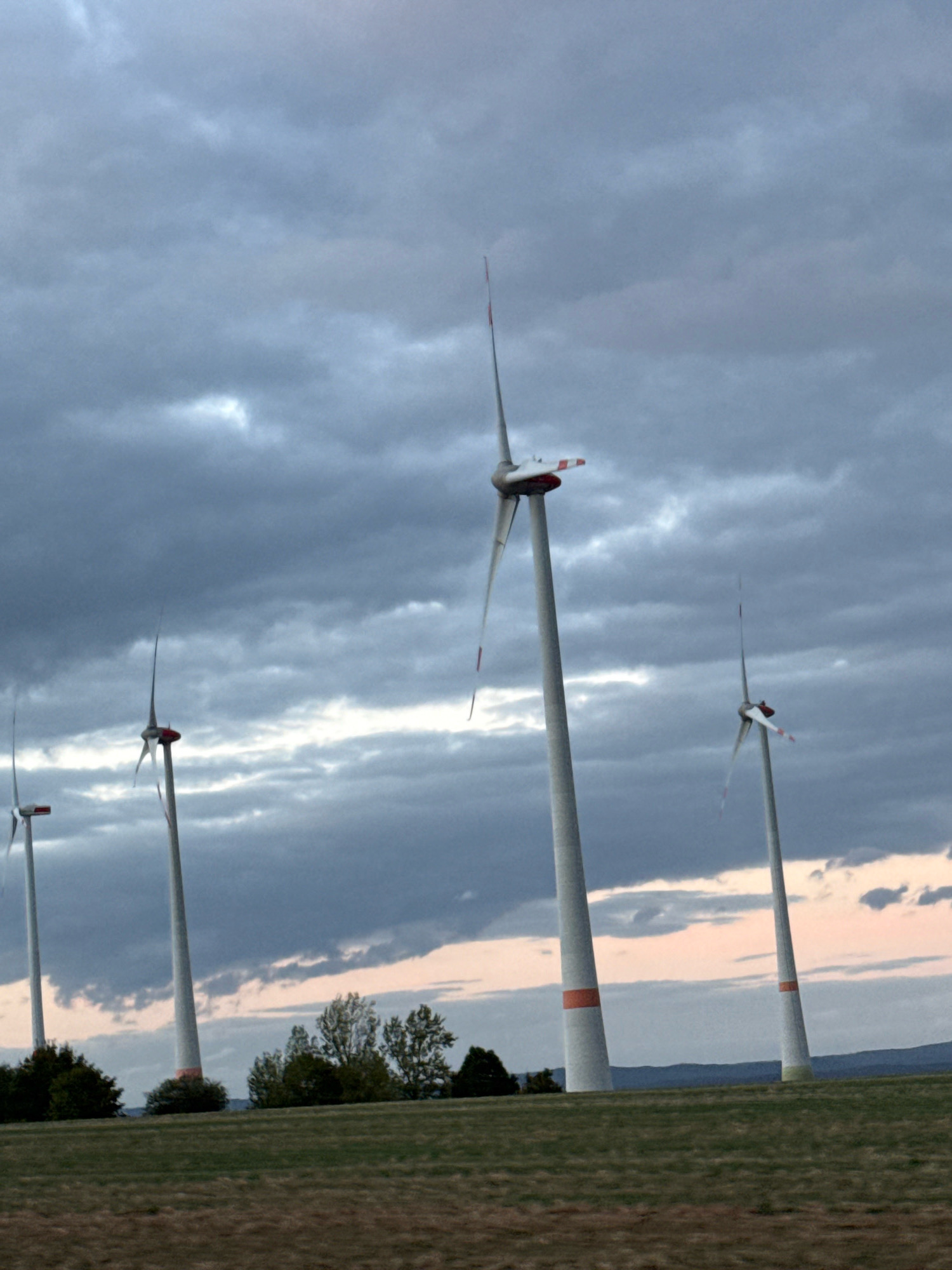 The image shows a wind farm with multiple wind turbines under a cloudy sky. The turbines are tall and white with red accents on their blades and bases. They are positioned in a grassy field with some trees in the background. The sky is filled with dark, dramatic clouds, suggesting a potentially stormy or overcast day. The overall scene depicts a landscape dotted with modern renewable energy infrastructure. The location of Sondershausen, Germany is not explicitly visible in the image but is provided as context for the scene.