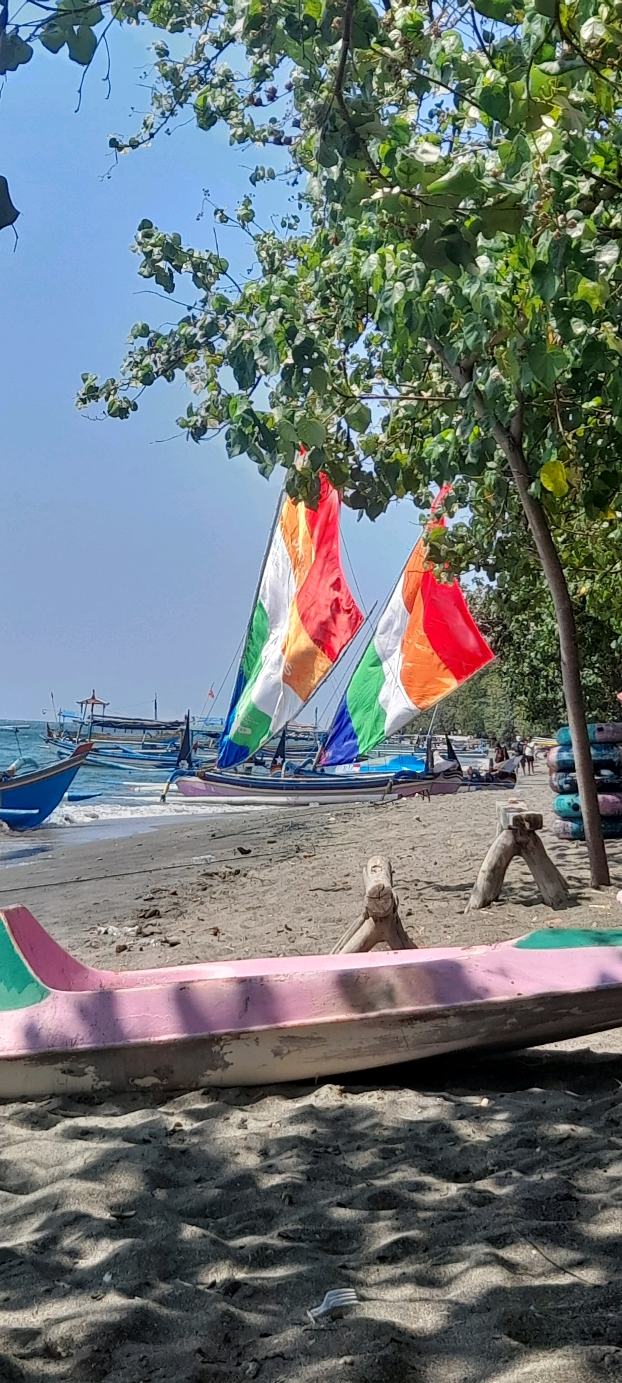 The image depicts a beach scene at Pasir Putih, Indonesia, under a clear blue sky. In the foreground, a pink and green boat lies on the sand. Behind it, the beach is sandy with some debris. Several other boats are anchored in the shallow water, with some featuring colorful sails in red, orange, green, and white. Trees with green foliage overhang the scene from the right. In the background, more boats are visible, and a few people can be seen walking near the water's edge. The overall environment suggests a sunny day at a coastal location.