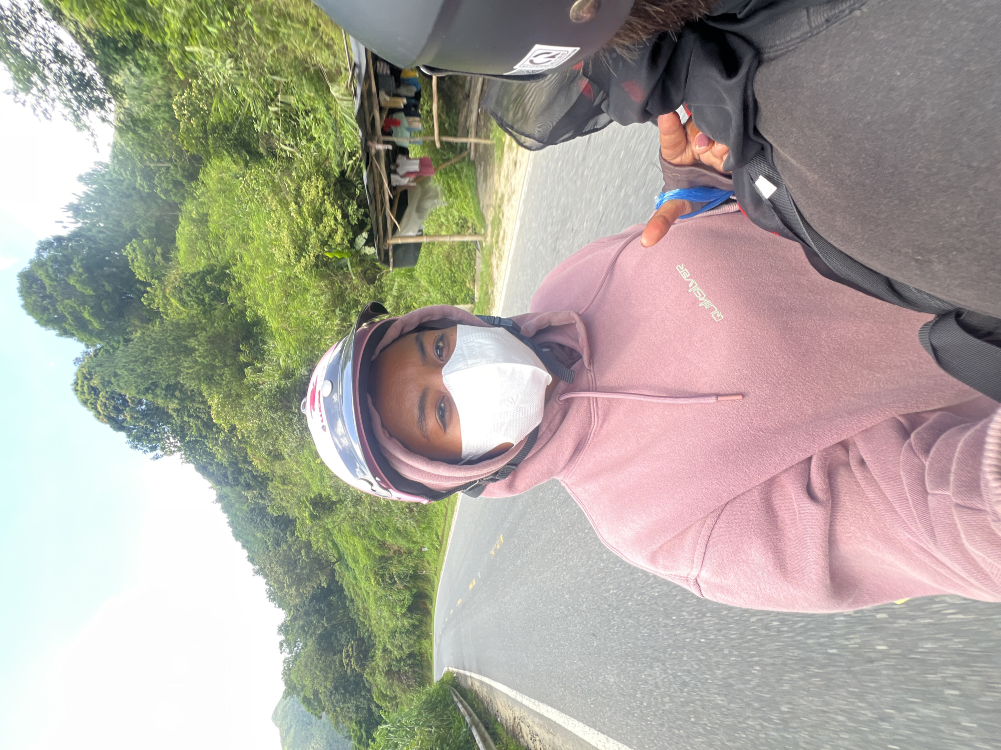 Two individuals are riding on a motorcycle on a paved road in a rural mountainous area, likely in Bình Lư, Vietnam. The person in the foreground, wearing a pink hoodie and a white face mask, is looking at the camera. They have a pink and white helmet on. Behind them, another person, wearing a black helmet, is visible from the back. The road curves to the left, bordered by a guardrail on the left and lush green vegetation and trees on the right. A small, makeshift shelter with clothes hanging on it is visible on the right side of the road. The sky is bright and clear with some clouds. The overall scene depicts a journey through a scenic, natural landscape.