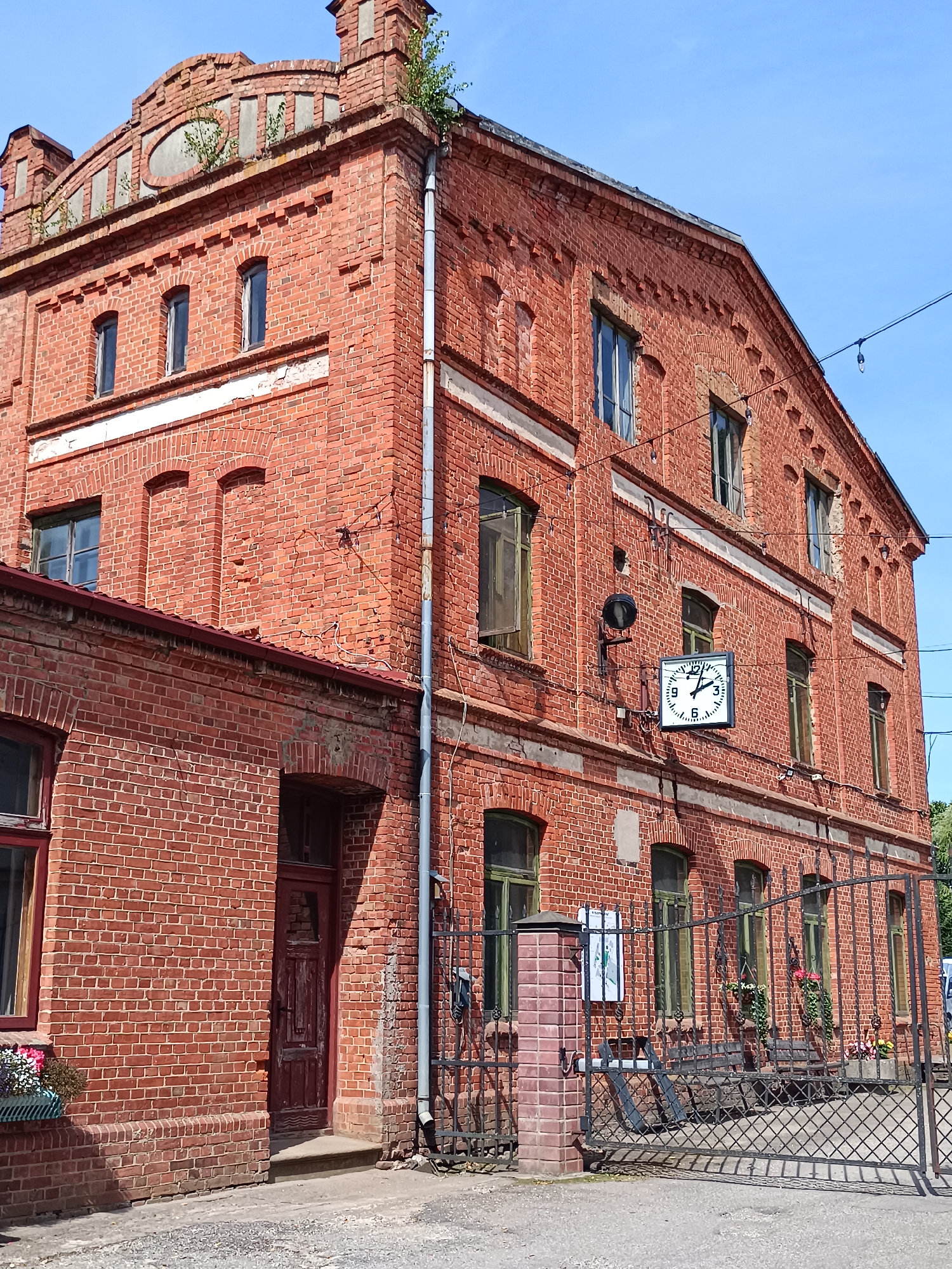 The image shows a red brick building in Aizpute, Latvia. The building appears to be old, with visible signs of age and wear. It has a distinctive architectural style with arched windows and decorative brickwork. A large clock is mounted on the exterior wall, showing the time as approximately 11:10. In the foreground, there is a metal gate and some benches, suggesting this might be a public space or part of a historical site. The sky is clear and blue. The overall scene evokes a sense of history and time.