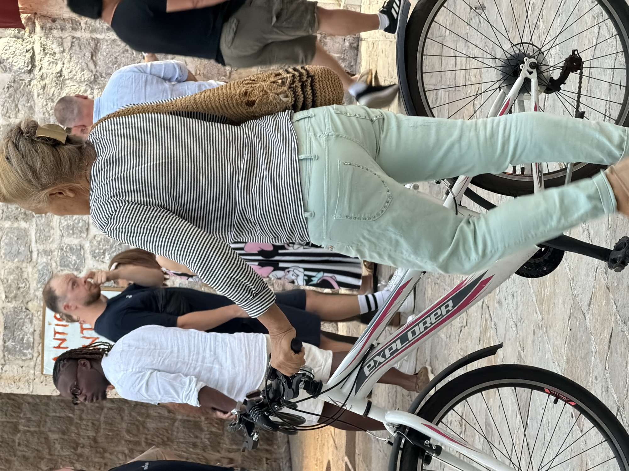 A woman with blonde hair pulled back, wearing a striped shirt and light green pants, stands next to a white bicycle with pink accents. She is holding the handlebars, with her right hand gripping the brake lever. The bicycle has the word "EXPLORER" printed on its frame. The background features a stone wall and several other people walking. The scene appears to be on a cobblestone street in Budva, Montenegro, likely near the Old Town walls, given the architecture. The overall impression is a casual daytime scene.