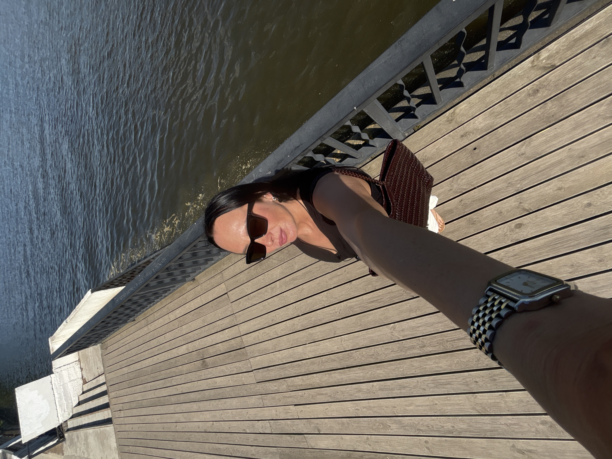 The image shows a woman on a wooden deck taking a selfie with water visible to the left. The setting appears to be in Chaikovsky, Russia, based on context clues (not visible in the image). The woman wears sunglasses and a black top and carries a woven brown bag. The scene includes a black metal railing, and the person taking the picture has a watch on their wrist.
