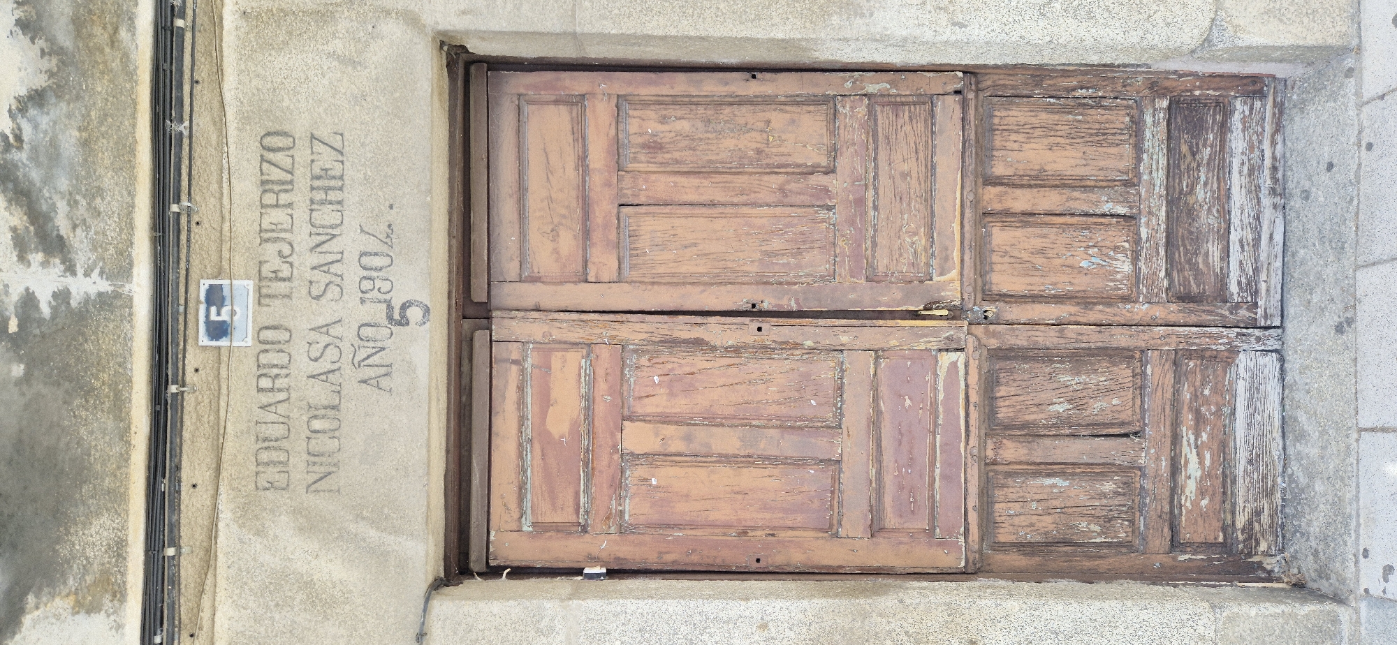The image shows a weathered, reddish-brown wooden double door set within a stone frame. The door is divided into four panels, with each panel featuring horizontal planks and a vertical element in the center. The wood is worn and shows signs of age, with peeling paint and exposed grain. The stone surrounding the door is textured and appears to be a light gray granite.
To the left of the door, etched into the stone, is text that appears to be names and a date. The text reads: "EDUARDO TEJERIZO NICOLASA SANCHEZ AÑO 1902. 5". Below the names and date, there is a small blue and white rectangular plaque with the number "5" inside it, attached to the stone wall with screws. There are also some black cables running vertically along the stone wall, secured with metal clips. The overall impression is one of age and history. The lighting suggests it is daytime, with no specific weather conditions apparent.
