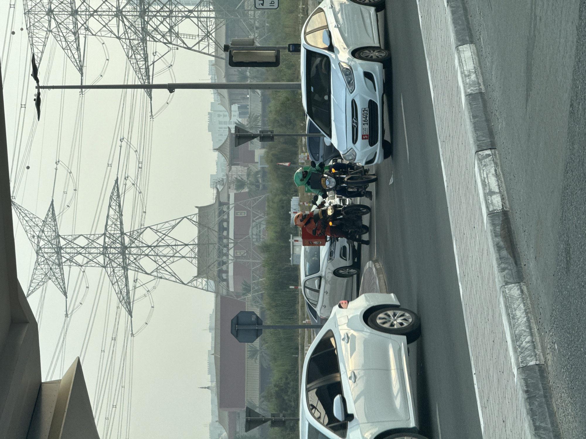 The image captures an outdoor urban scene, likely in Dubai, United Arab Emirates, characterized by its hazy atmosphere and prominent high-voltage power line towers. In the foreground, two white sedans are on a paved road, one on the left and another slightly behind and to the right. The sedan on the right has a license plate that reads "5 16401". Between the two cars, two individuals on motorcycles are visible. The rider on the left wears an orange helmet and a red top, while the rider on the right wears a green helmet and a green jacket, both appearing to be delivery personnel. They are stopped at what appears to be a traffic light or intersection. In the background, several buildings with distinctive East Asian architectural influences, including curved roofs, are present, suggesting a themed area or development. Tall, lattice-like power line towers dominate the sky, indicating proximity to major electrical infrastructure. The overall environment suggests a warm climate with a noticeable haze.
