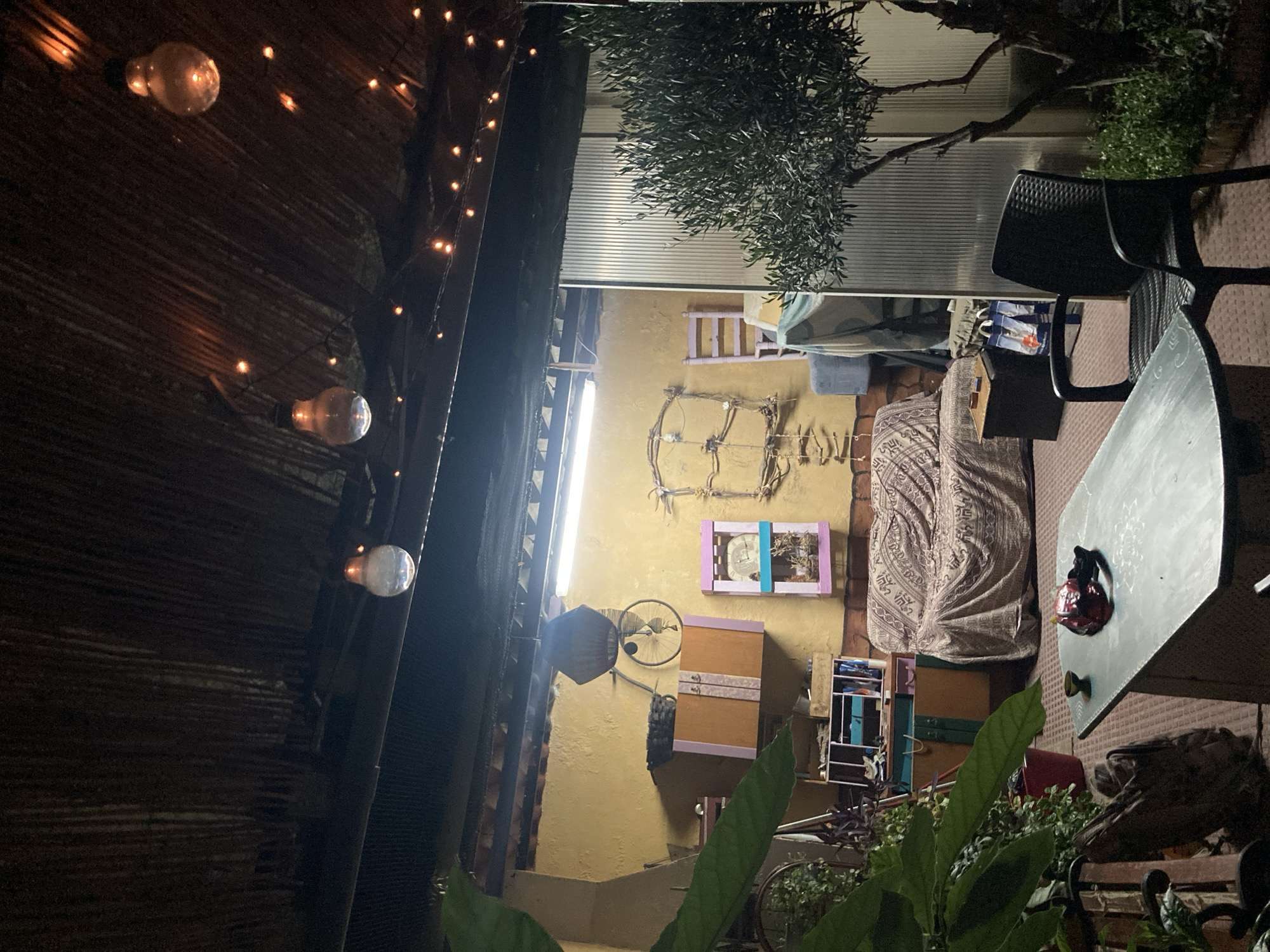 This is an outdoor patio scene at night, with a corrugated roof overhead. String lights and three exposed light bulbs are visible, illuminating the space. On the wall, there is a yellow-painted section with various objects, including a bicycle wheel with a basket attached, a cabinet, a decorative frame made of branches, and a clock in a pink frame. A sofa covered with a patterned fabric is centrally located, with a small wooden side table next to it. To the right, an olive tree stands with dense foliage, and behind it, a corrugated plastic wall. In the foreground, a table with a light-colored surface holds a small dark bowl and a red object. A black plastic chair is positioned to the right of the table. The ground is covered with a patterned rug. The scene appears to be a relaxed outdoor living area, possibly a backyard or patio of a residence in Sabadell, Spain.