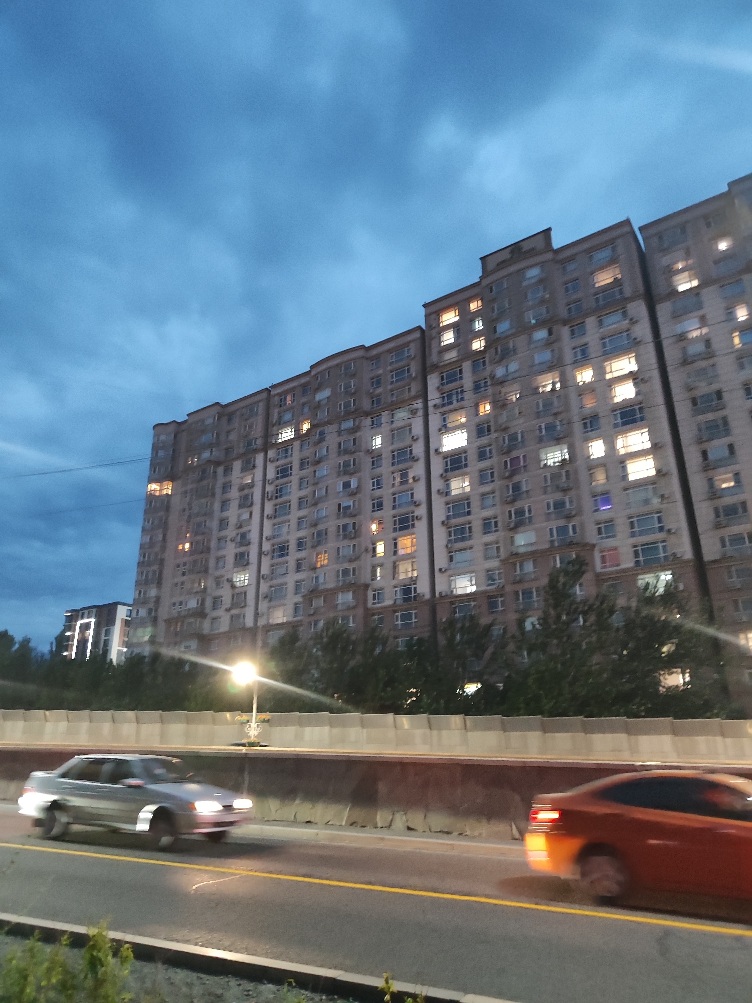 The image depicts a twilight scene in Almaty, Kazakhstan, featuring a long, multi-story apartment building with numerous illuminated windows. In the foreground, two cars are visible on a road; a silver sedan is in motion on the left, and an orange coupe is moving to the right on the right side of the frame. Trees and a concrete barrier line the road. The sky is overcast with a deep blue hue. The apartment building appears to be composed of multiple connected sections, with varying architectural details and window designs.