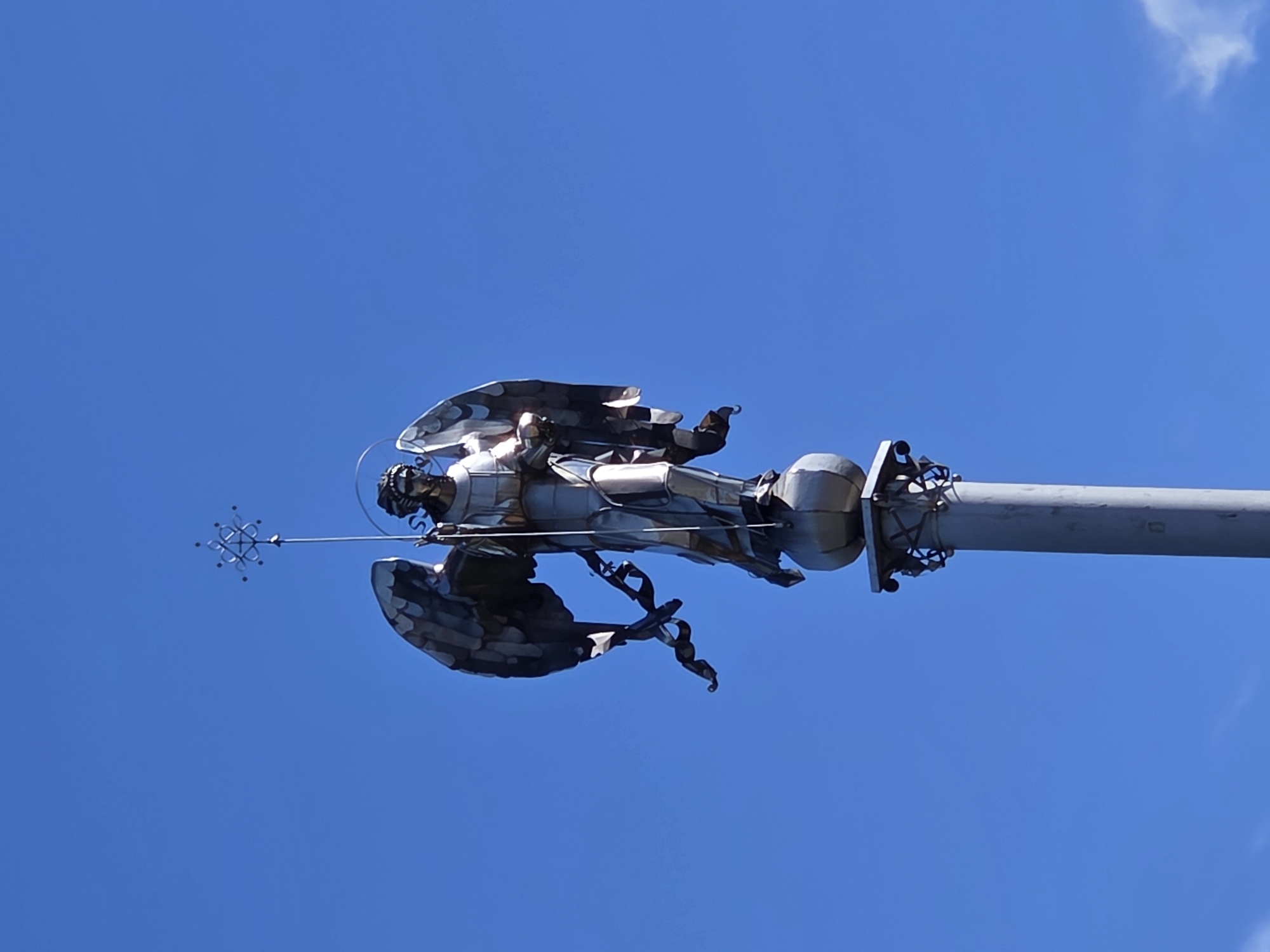 The image shows a statue of an angel atop a column against a clear blue sky. The angel is depicted with large wings, a halo, and is holding a staff topped with a decorative element. The statue appears to be made of metal and has a polished, reflective surface. The column is cylindrical and grey, with decorative elements near its top. The overall scene suggests a monument or a religious statue located in an outdoor public space. While the image itself does not provide specific location details, if this statue is indeed located in Podolsk, Russia, it is likely a significant landmark within the city.