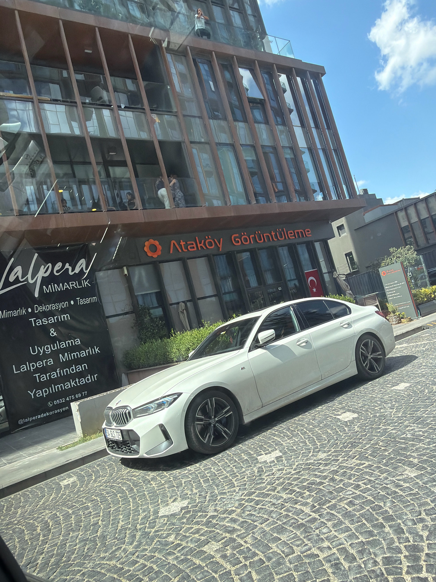 In Bakırköy, Turkey, a white sedan is parked on a cobblestone street in front of a modern building with a glass facade. The building features horizontal wooden beams and multiple stories. A large black banner with white text is displayed on the side of the building, advertising "Lalpera Mimarlık" which offers architectural design, decoration, and application services. Above the banner, the text "Ataköy Görüntüleme" is visible in orange lettering. A Turkish flag is displayed on a pole near the building entrance. People are visible on the upper floors of the building, some looking out of windows, and one individual is standing on a balcony. The sky is blue with scattered white clouds.