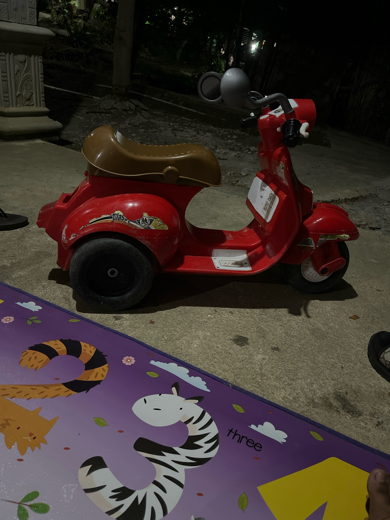 A red toy scooter, resembling a Vespa, is positioned on a concrete surface at night. The scooter has a brown seat and a silver rearview mirror. A purple mat with cartoon animal illustrations and the word "three" is in the foreground. The background shows trees and foliage, suggesting an outdoor, possibly residential, setting. The image was taken in Wonokerto, Indonesia.