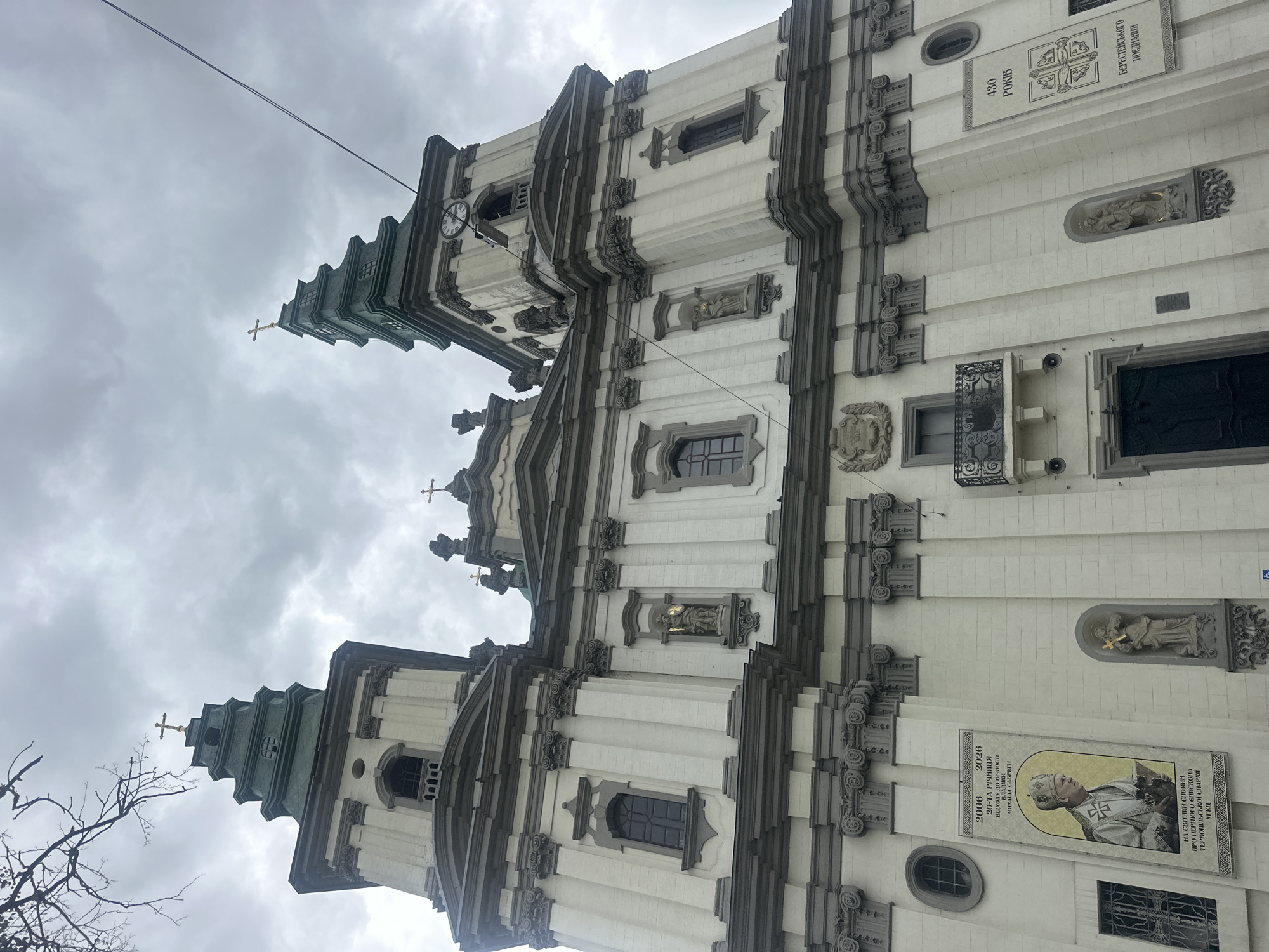 The image shows the Ternopil Cathedral in Ternopil, Ukraine, on a cloudy day. The cathedral is a large, imposing structure with two tall towers topped with green spires and crosses. The facade is made of white stone with gray architectural details, including columns, arches, and sculptures. There are several windows and niches on the facade, some containing statues of religious figures.
On the left side of the facade, there is a large banner with a portrait of a man in religious attire, along with text in Ukrainian. The text appears to commemorate an anniversary. Below this banner is a window with decorative iron bars.
On the right side of the facade, there is another banner with text and a symbol, also in Ukrainian. There is a clock visible on the right tower. A balcony with ornate iron railings juts out from the facade above a large, dark doorway. The sky is overcast with heavy gray clouds.