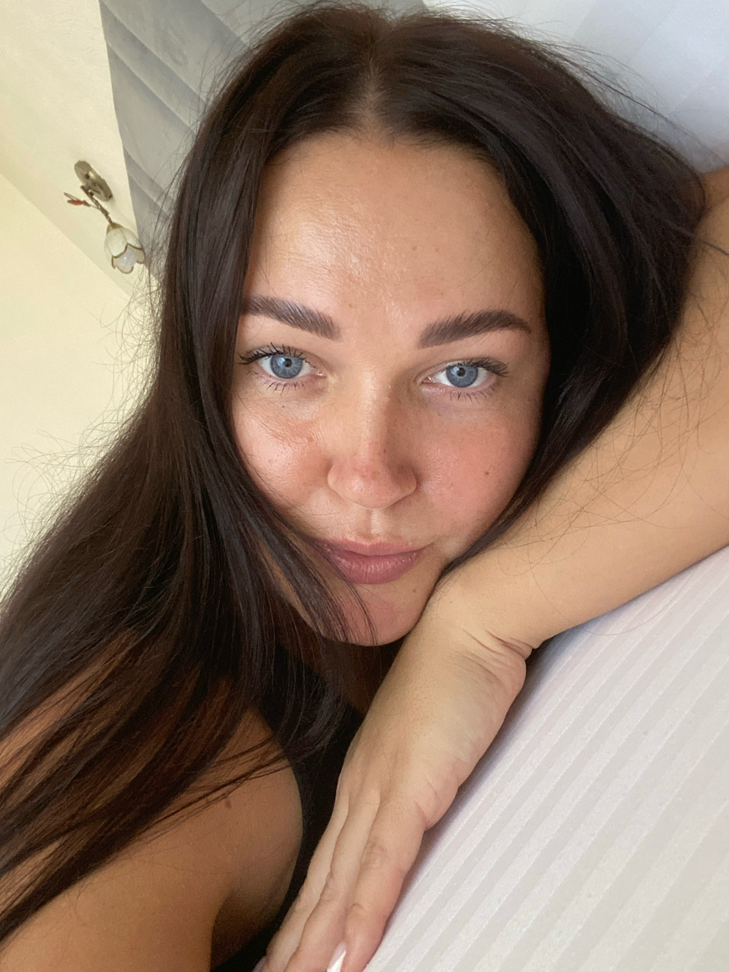 A close-up, head and shoulder shot features a woman with dark brown hair and blue eyes, lying on her side and resting her head on her arm. The background consists of a light-colored wall with a decorative light fixture on the left and a white, striped surface, likely a bedspread, on the right. The lighting suggests an indoor setting, possibly a bedroom. The woman is looking directly at the camera with a neutral expression. There is no indication of the city of Nebug, Russia within the image itself.