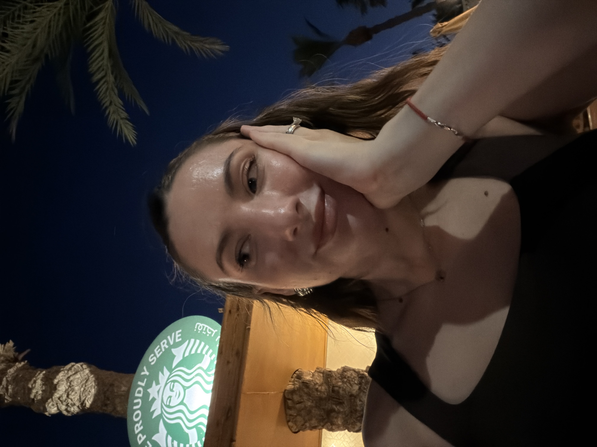 The image is a selfie taken at night in Sharm El Sheikh, Egypt, likely outdoors. The subject is a young woman with long, wavy brown hair. She is wearing a black tank top and a delicate necklace. Her right hand is on her cheek, and she is wearing a ring on her left hand, which is partially visible. A red bracelet is also visible on her wrist.
In the background, a palm tree is silhouetted against a dark blue night sky. To the left, a round, green Starbucks logo is illuminated, with the text "PROUDLY SERVE" and the Starbucks siren visible. The sign is attached to a wooden structure. The overall lighting suggests it is nighttime, possibly illuminated by artificial lights from the Starbucks establishment. The woman's expression appears relaxed and happy, indicating a pleasant social or personal moment.
