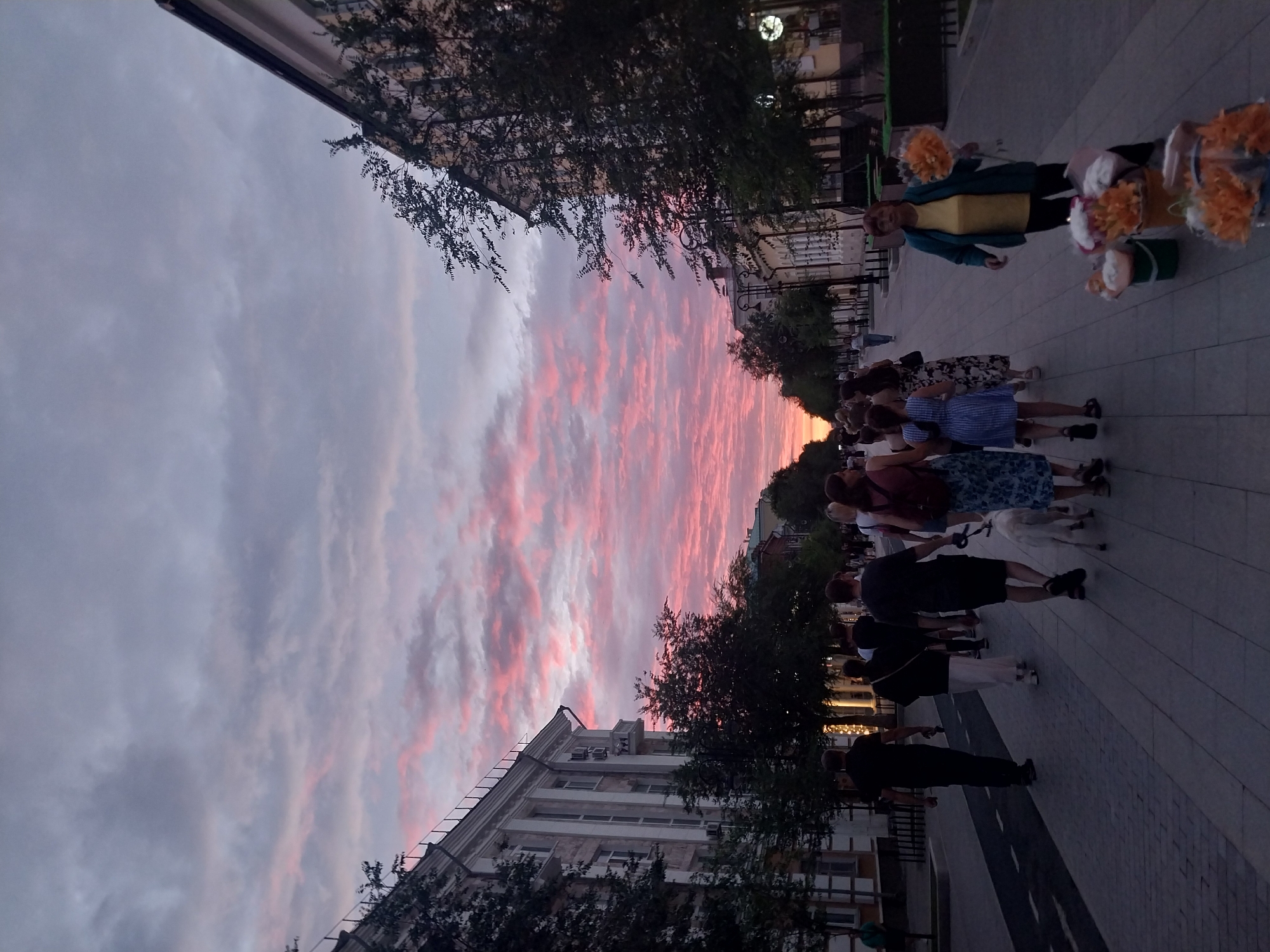 The image depicts a street scene in Orenburg, Russia, at sunset. The sky is filled with dramatic pink and grey clouds. On the left side of the street, a large, ornate building with a white facade and classical columns lines the sidewalk. Trees with dark green foliage are planted along the street, casting shadows. Several people are walking along the sidewalk, mostly in silhouette. A woman is selling flowers from buckets on the right side of the street. The pavement is made of grey bricks. The overall impression is of a calm evening stroll on a city street during a vibrant sunset.
