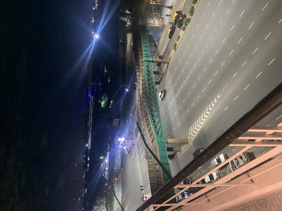 An elevated view at night reveals a mostly deserted city street divided by dashed lines and bordered by a green metal structure. Cars and scooters are parked along the edges of the street, while a few vehicles move along the empty lanes. Streetlights illuminate the scene, casting a bright glow on the surrounding buildings and infrastructure. The photograph is taken from a high vantage point, looking down on the urban landscape.