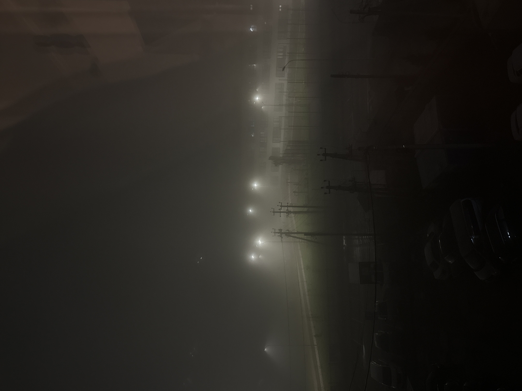 A nighttime scene depicts a city obscured by heavy fog. Streetlights illuminate the fog, creating a diffused glow. Power lines and poles are visible in the midground, partially obscured by the fog. Cars are parked along the street on the right side of the image. A large building is visible in the background behind the fog.