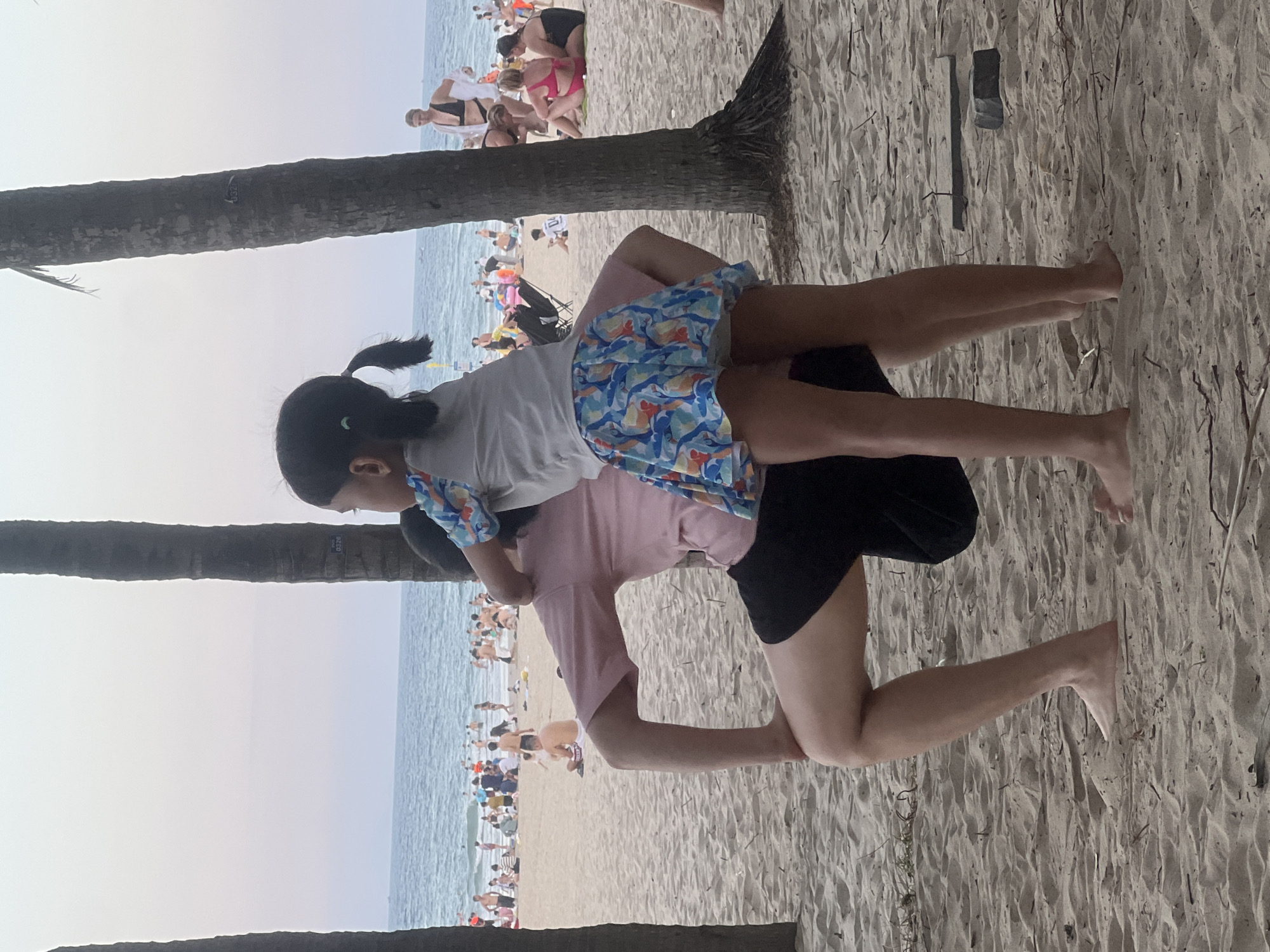 A young girl with dark hair in pigtails stands on the shoulders of an adult who is crouching on a sandy beach, likely at My Khe Beach in Đà Nẵng, Vietnam, given the presence of palm trees and a crowded oceanfront. The girl is wearing a light gray t-shirt with a colorful patterned skirt featuring blue, orange, and white fish. The adult is wearing a pink t-shirt and black shorts. In the background, the ocean is visible with numerous people swimming and sunbathing. Other palm trees and beachgoers are scattered across the sand. The overall scene depicts a recreational moment on a crowded beach.
