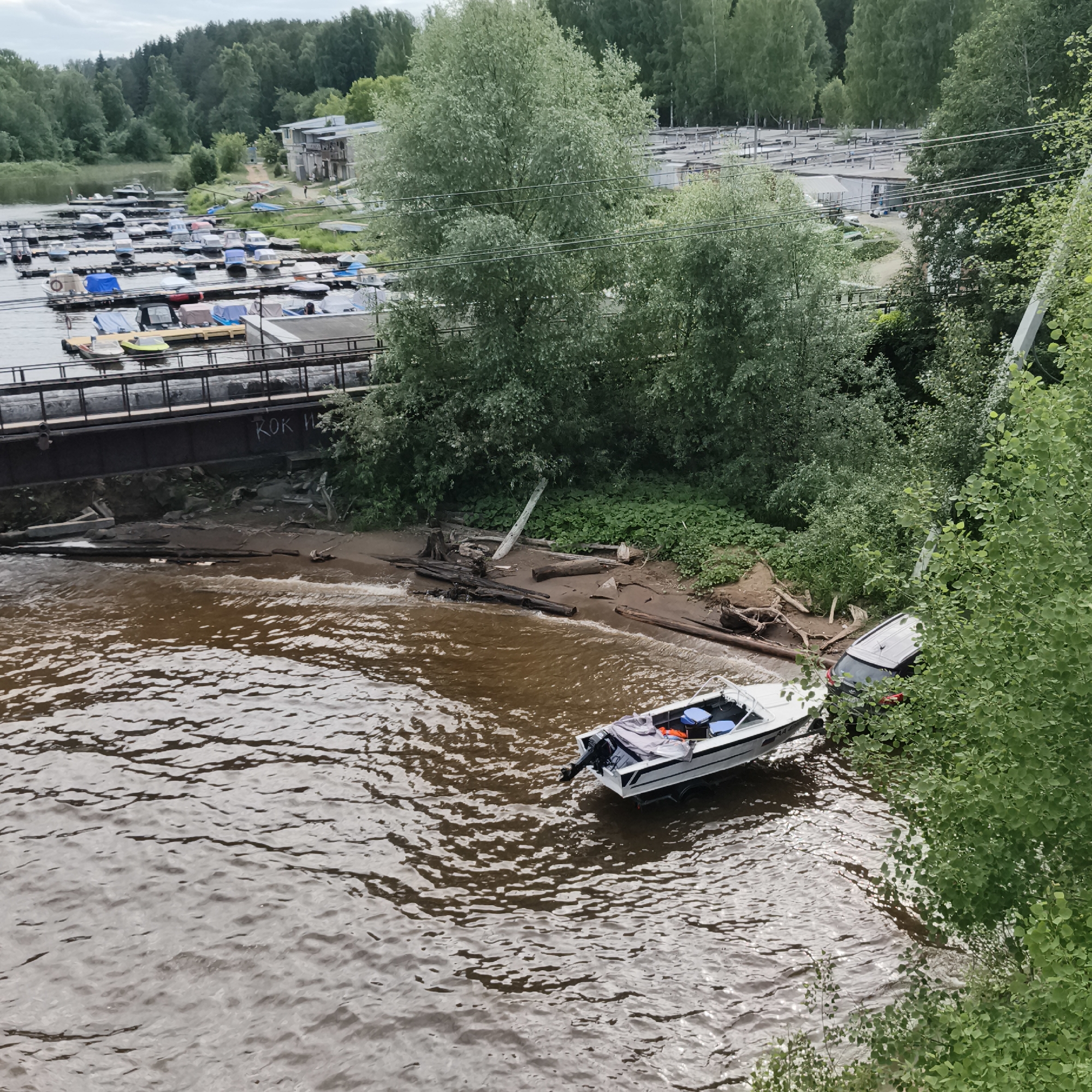 The image shows a riverbank with a boat dock, several boats, and a bridge. The river is brown and appears to be calm. The riverbank is lined with trees, and there are some debris scattered along the shore. A white speed boat is partially in the water and partially on the bank, with a grey car also partially submerged in the water next to it. Several other boats are docked in the background, some covered with tarps. A bridge crosses the river in the foreground, and graffiti is visible on its side. The sky is overcast, suggesting a cloudy day. The scene appears to be in a rural or semi-rural setting, possibly a recreational area. The presence of boats and a dock indicates a location near a body of water, suitable for boating and water sports. The car partially submerged in the water suggests a recent accident or flooding. The graffiti on the bridge reads "ROK H".