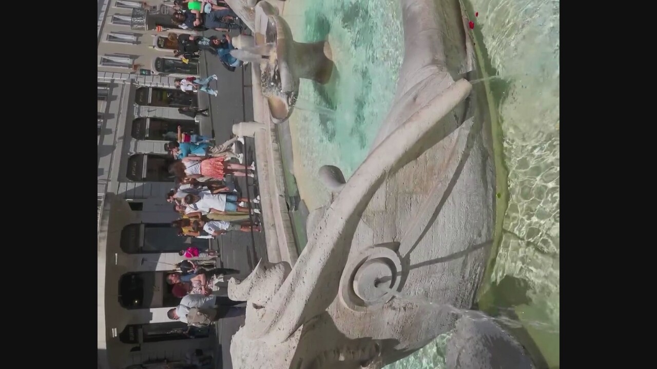 A bright, sunny day shows a public fountain with a stone carving of a fish head, its mouth open to allow water to flow into the pool. In the background, a street is lined with shops and a crowd of tourists. Many people are walking or standing around, some taking pictures.