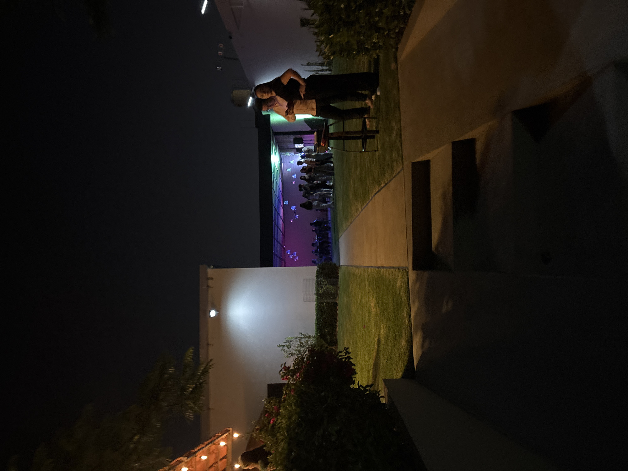 Captured at night in San Bernardino Tlaxcalancingo, Mexico, the image shows an outdoor event. A group of people stands under colorful lights, with two individuals closer to the viewer standing on a pathway alongside green foliage. The scene is illuminated by outdoor lighting and string lights in the distance.