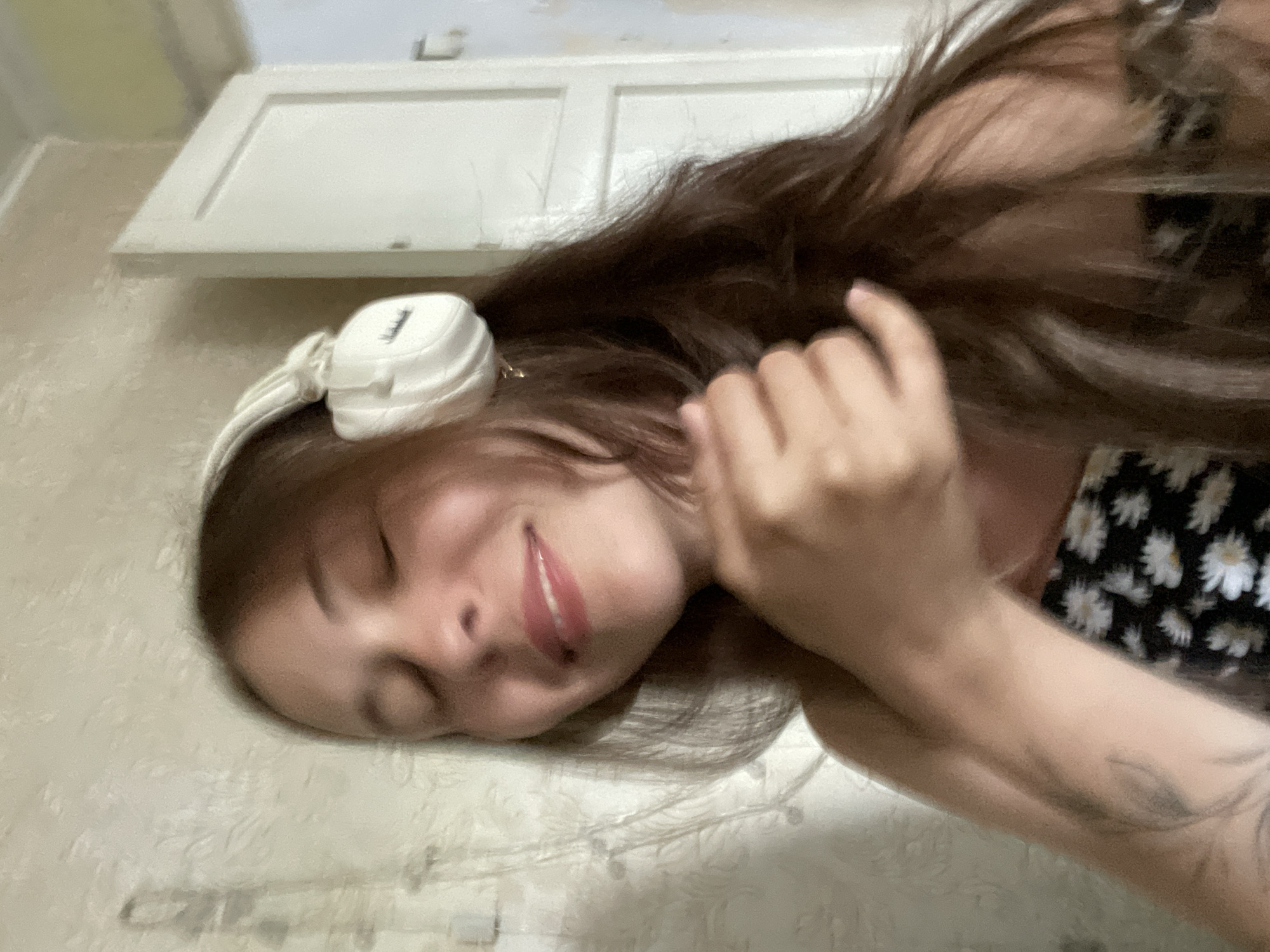 A woman with long, dark hair and closed eyes smiles while wearing white headphones. She is wearing a black and white floral top. Her right hand is resting on her chest. A tattoo is visible on her left arm. The background appears to be an indoor setting with a textured wall and a white door.