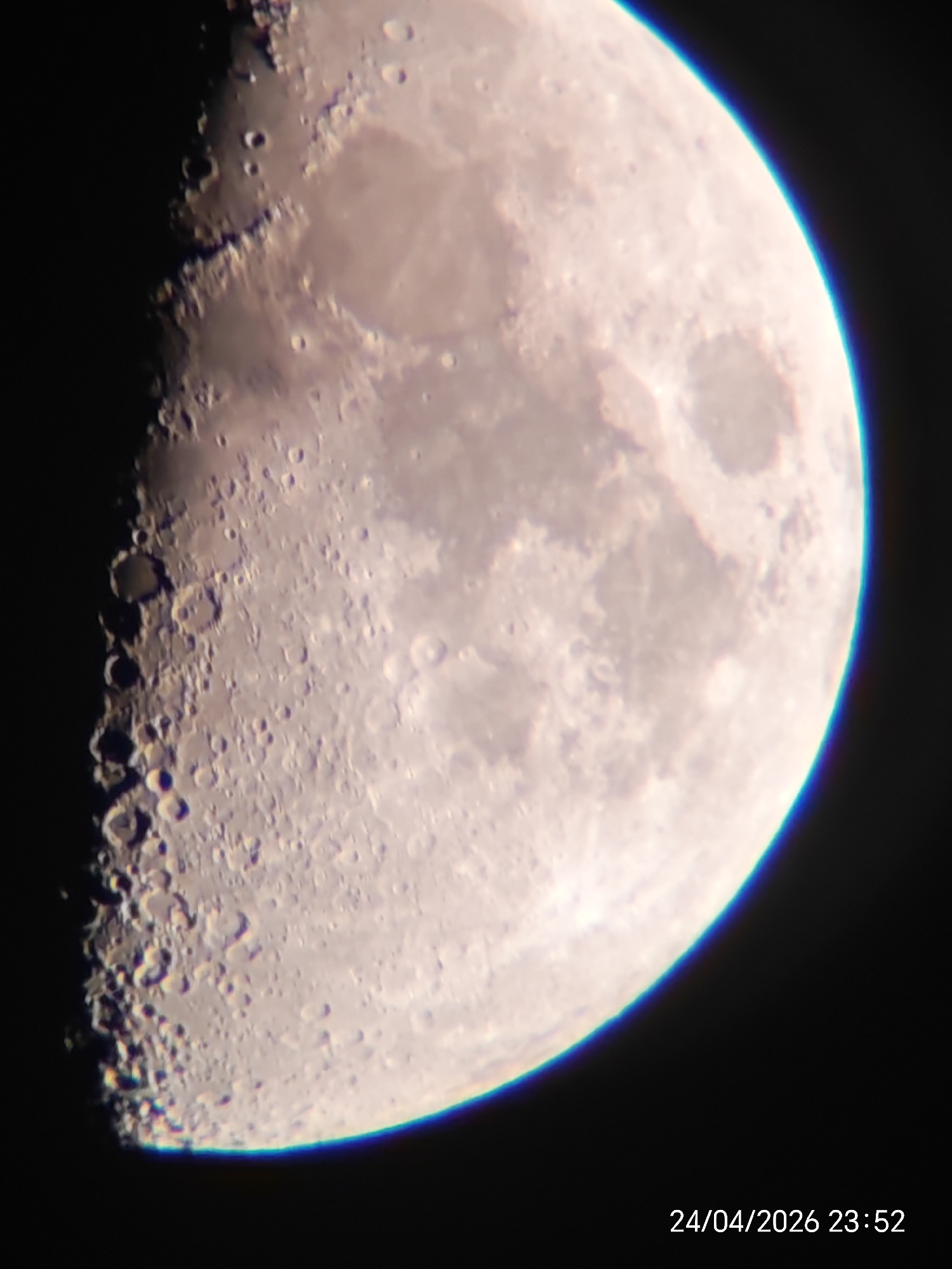 This is a detailed, close-up photograph of the Moon, specifically depicting a waxing gibbous phase. The Moon occupies the majority of the frame, set against a deep black background of space.
The right side of the Moon is brightly illuminated, curving gently into the darkness, while the left side shows a distinct terminator line where light transitions to shadow. Along this terminator, numerous craters are visible, particularly in the lower-left quadrant, appearing rugged and casting prominent shadows that highlight their depth and structure. Further into the illuminated portion, vast, smoother, darker plains (maria) are visible, alongside scattered smaller craters and lighter, mountainous regions.
A notable detail is the slight blue and purple chromatic aberration visible along the brightly lit edge of the Moon, especially towards the top and bottom right, indicating an optical artifact from the lens or telescope used. The overall clarity of the image allows for good appreciation of the lunar surface features.
The image captures an instance of astrophotography or lunar observation, showcasing the Moon's intricate topography under nocturnal conditions. In the bottom right corner of the original image, white text displays the date and time: "24/04/2026 23:52".