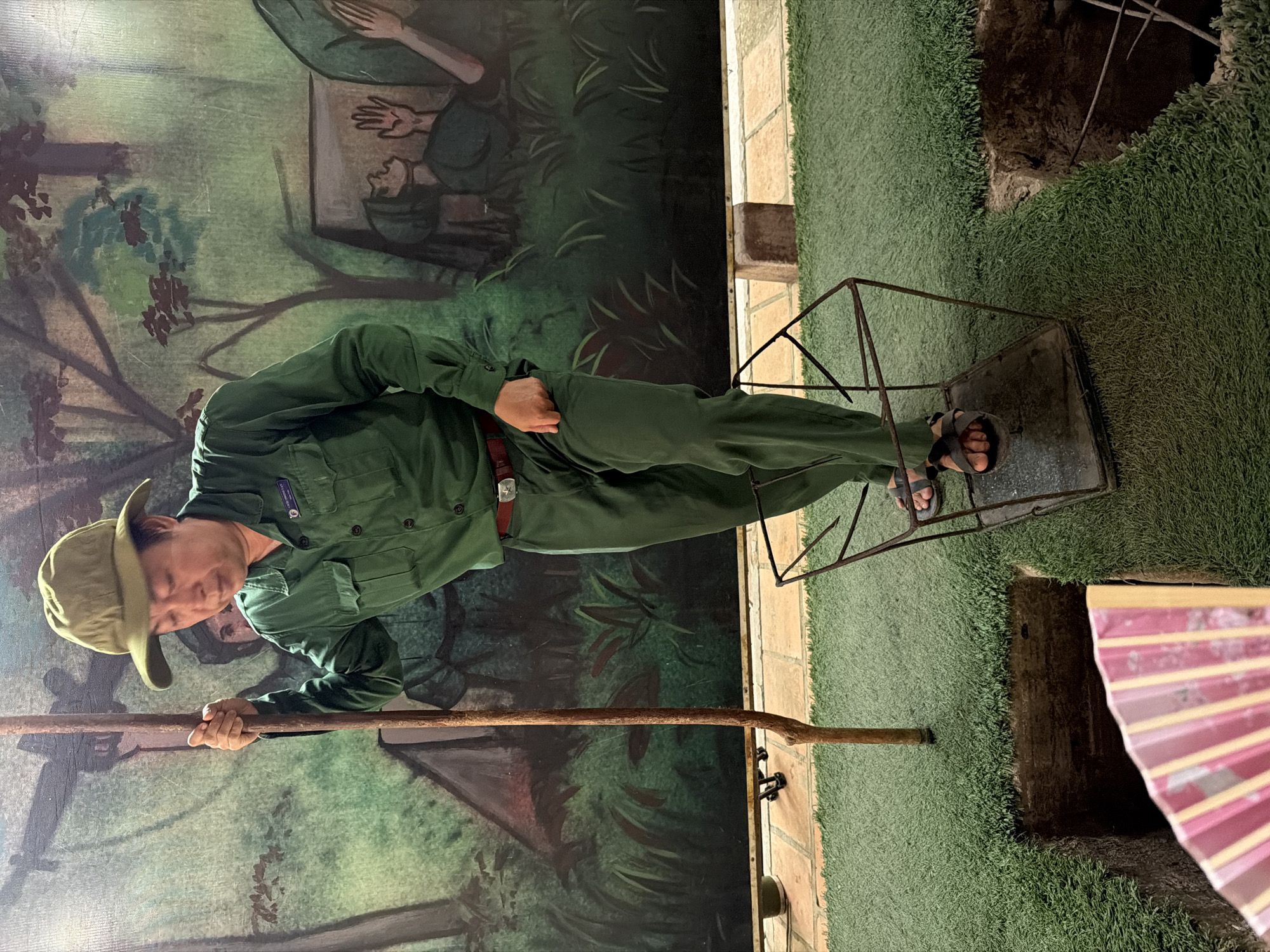 This media file captures an indoor exhibit, likely a diorama, focused on the Cu Chi Tunnels near Ho Chi Minh City, Vietnam. The central subject is a person, possibly a demonstrator or mannequin, dressed in a green military-style uniform, including a shirt, trousers, and a wide-brimmed hat. They wear sandals and a brown belt with a metallic buckle. The person is standing upright, holding a long wooden staff with both hands, positioned vertically in front of them.
Around the person's feet is an open, rusted metal frame, resembling a narrow cage, which appears to be placed on artificial green turf that covers the ground. To the right of the person, a dark, narrow hole in the ground is visible, suggesting an entrance to a tunnel system. A portion of a pink and yellow handheld fan is also visible in the bottom right corner.
The backdrop is a painted mural depicting a jungle or forest scene with various shades of green, brown, and hints of blue, showing trees, foliage, and stylized figures of soldiers in combat or camouflage. The overall activity depicted is a demonstration or re-enactment of navigating or illustrating the conditions of the Cu Chi Tunnels.
Notable details include the words "CỦ CHI VIỆT NAM" clearly visible on the left pocket of the person's uniform, explicitly identifying the exhibit's theme and location. The metal frame around the feet typically represents the constrained dimensions of the tunnels. The setting is indoors with artificial lighting, and there are no discernible weather cues. The scene aims to be informative and historical.