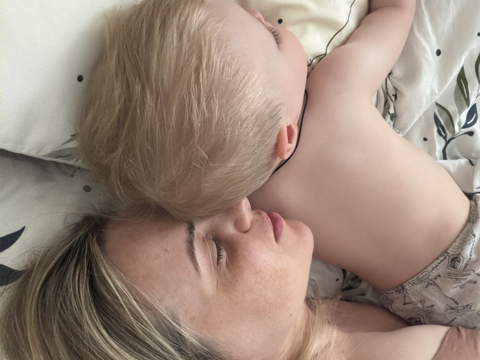 A woman and a young child are lying down. The child's head rests on the woman's head. The woman is fair-skinned with blonde hair, closed eyes, and freckles. The child has fair skin and blonde hair. They are lying on a bed with a patterned sheet and pillow. There is no information available to determine if the image was taken in Votkinsk, Russia.