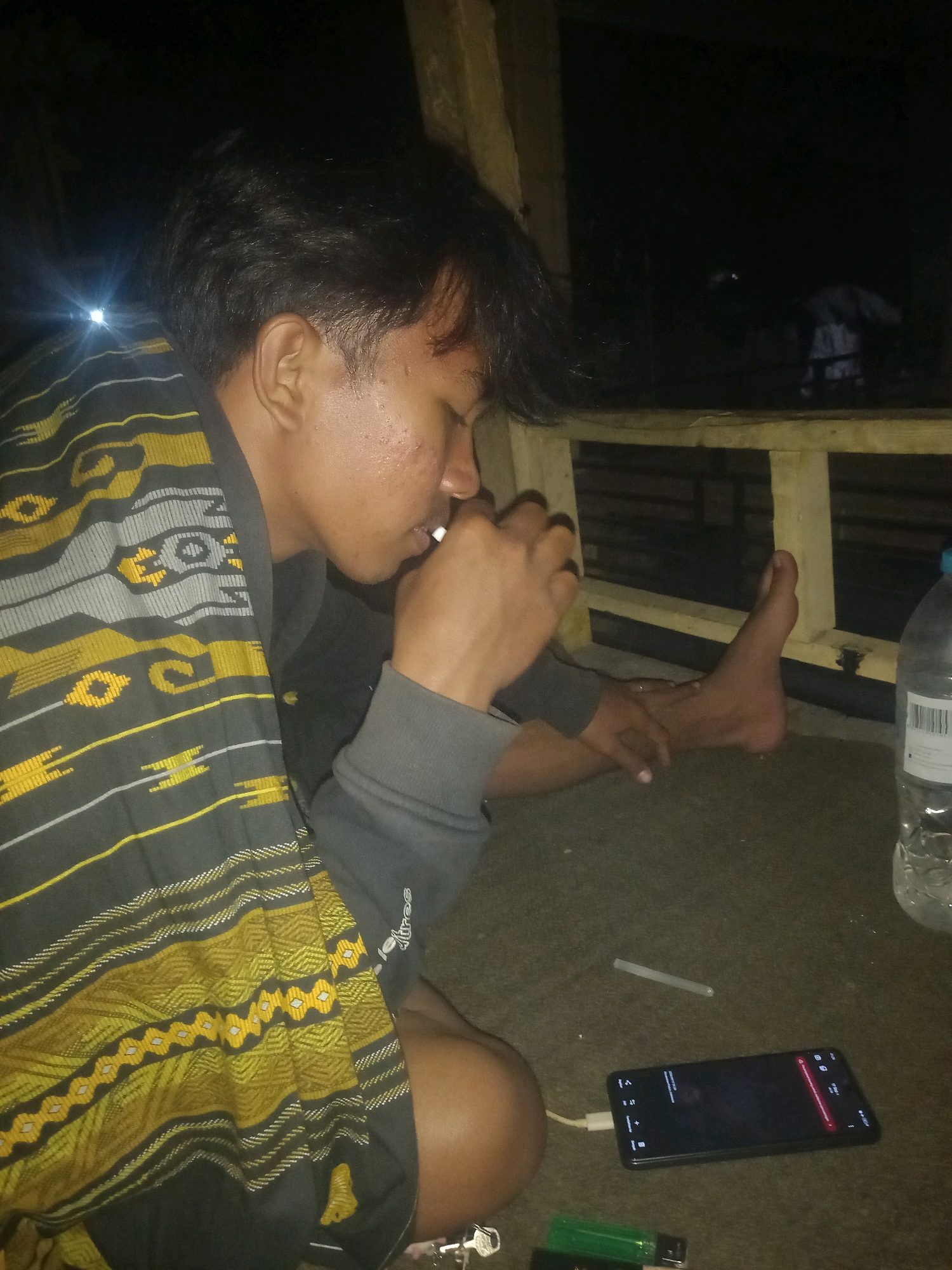 In a dimly lit outdoor setting, likely in Paiton, Indonesia, a young man is shown from the chest up. He is seated and wearing a dark grey hoodie over a patterned sarong with intricate yellow and grey designs. His right hand is raised to his mouth, holding a lit cigarette. His eyes are closed, and his head is slightly tilted downwards. To his right, a smartphone is placed on a dark surface, displaying a red notification or alert. Adjacent to the phone are a green lighter and a thin white object, possibly a cigarette filter or a glow stick. In the background, a wooden structure and a plastic water bottle are visible, with darkness beyond.