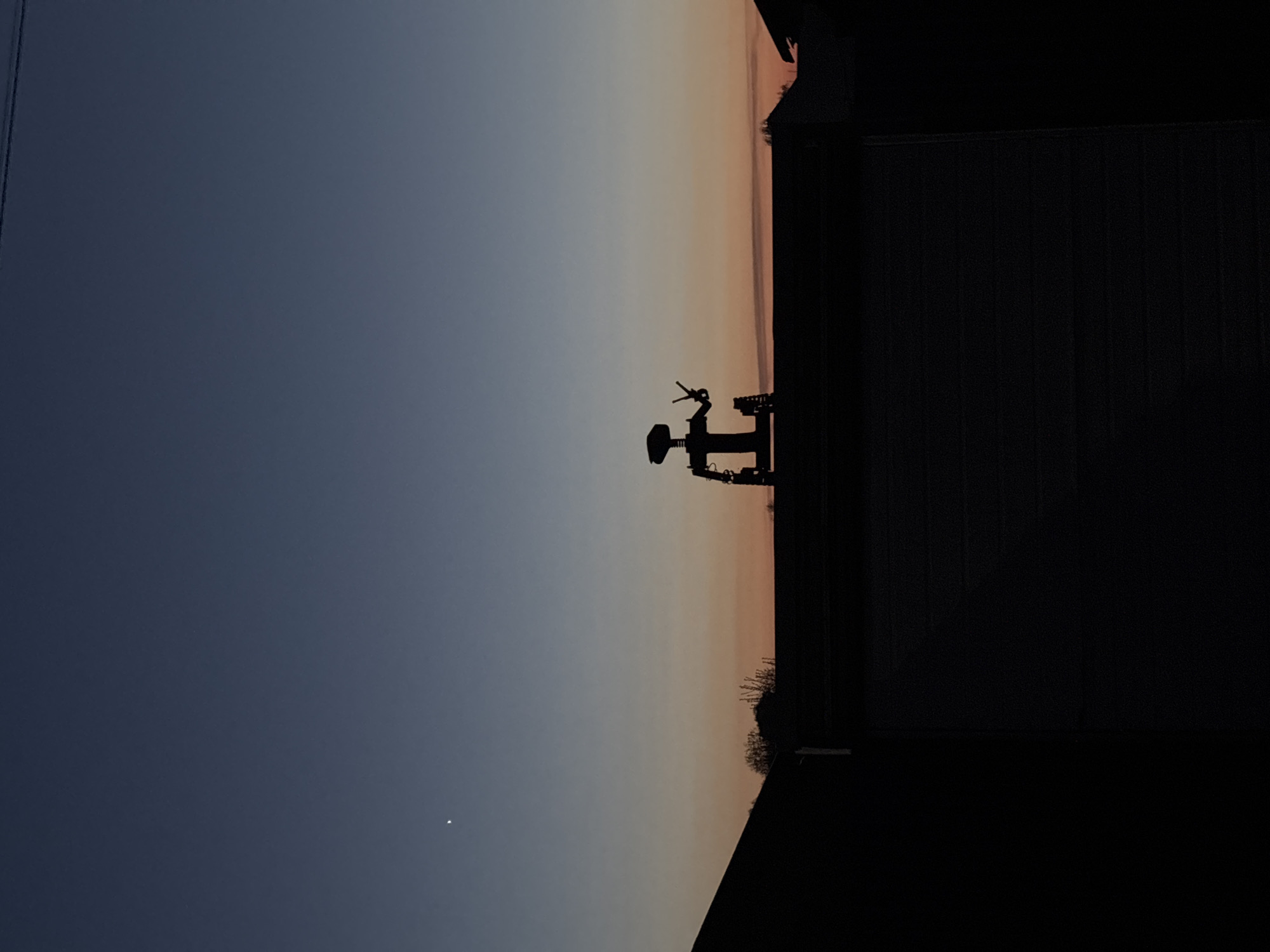 The image shows a silhouette of an object resembling a water tower or some kind of machinery, located on a surface that transitions into a dark area. Behind the object, there is a gradient sky ranging from light orange near the horizon to a blue hue. A faint point of light is visible in the lower left portion of the sky, likely a star or a distant light source.
I do not have enough information to determine the location in Фоки, Russia.