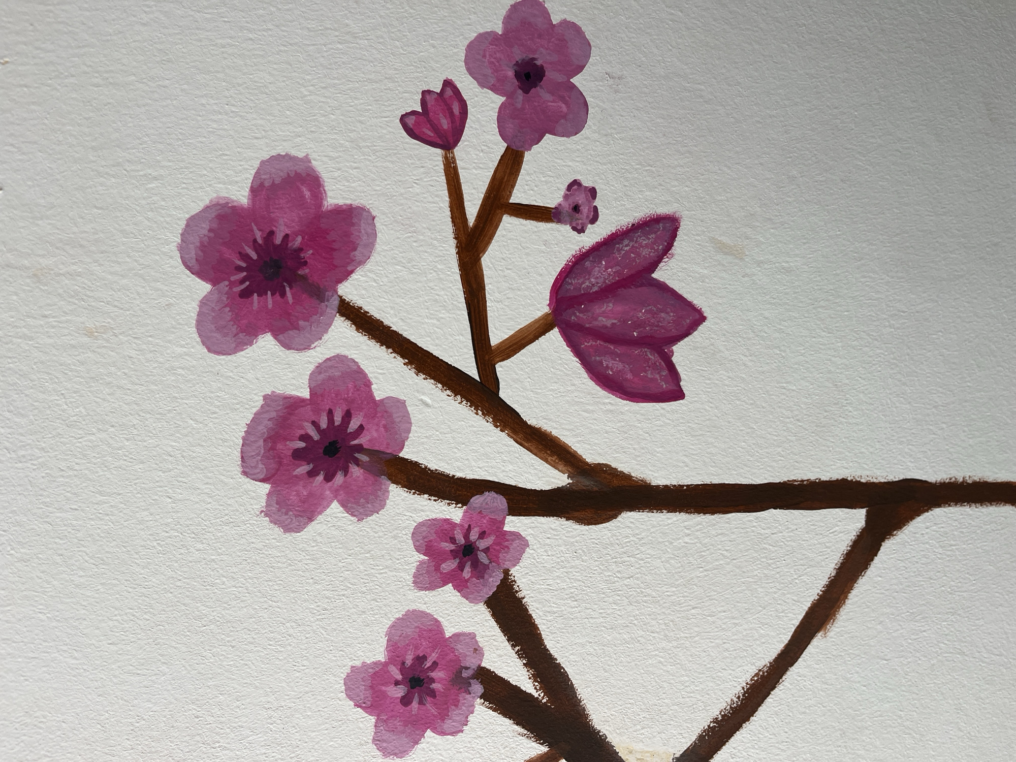 The image presents a close-up view of a painting depicting a stylized branch with pink flowers and buds, set against a textured, off-white background. The scene focuses on a brown branch, rendered with visible brushstrokes, which extends diagonally across the frame.
Several flowers in various stages of bloom are attached to the branch. The prominent blossoms are painted in shades of pink and magenta, featuring five petals and darker, purplish-pink centers. Examples include a large open flower on the bottom left, another above it, and one near the top center. The painting also shows several buds: a well-defined magenta bud with closed petals near the top left, a smaller, more nascent bud further up the branch, and a larger, somewhat abstract magenta form on the right side which could be a developing flower or stylized leaves. The painting style is somewhat naive or folk-art inspired, utilizing opaque colors like acrylic or gouache.
The background is a textured, light-colored surface, possibly a wall or a canvas with a distinct grain, which adds depth to the simple floral design. The lighting is even, illuminating the painting without harsh shadows. No text is visible within the image. This artwork was photographed in Alanya, Turkey.