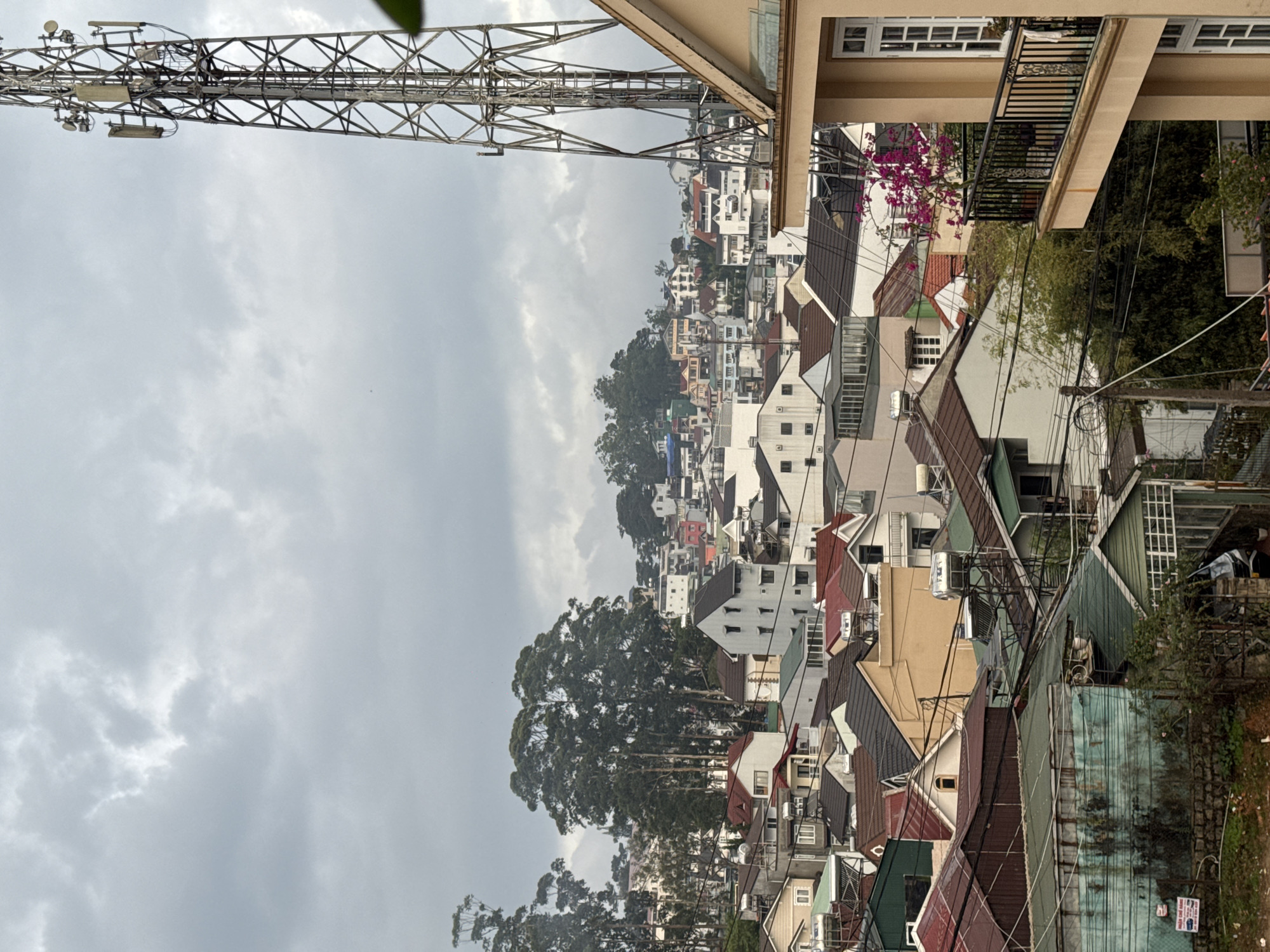 The image depicts a cityscape, possibly in Da Lat, Vietnam, based on the architectural style and general appearance. The view is from an elevated position, looking down over a dense arrangement of buildings, primarily houses with varying roof colors including red, brown, and green. A communication tower occupies the upper left portion of the frame. The sky is overcast with gray clouds. A building with a balcony is situated in the upper right corner. Numerous trees are scattered throughout the cityscape. There are some power lines running across the rooftops.
