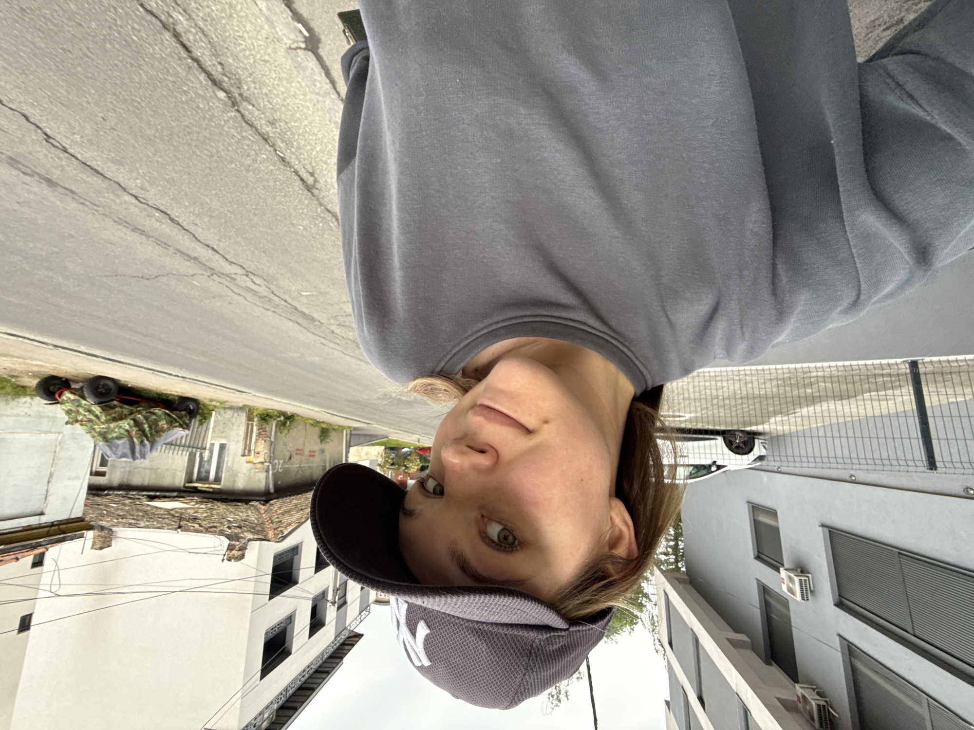 A young person wearing a gray sweatshirt and a gray New York Yankees baseball cap is the central subject. They are facing the camera with a slight smile. The background reveals an urban street scene in the Vozdovac Urban Municipality, Serbia. The street is cracked and aged. On the left, there are buildings, one with white walls and dark windows, and another slightly dilapidated with a tiled roof. A small, camouflage-covered vehicle is parked in front of these buildings. On the right is a modern building with a gray facade, windows, and air conditioning units. A white car is parked along the side of the street.