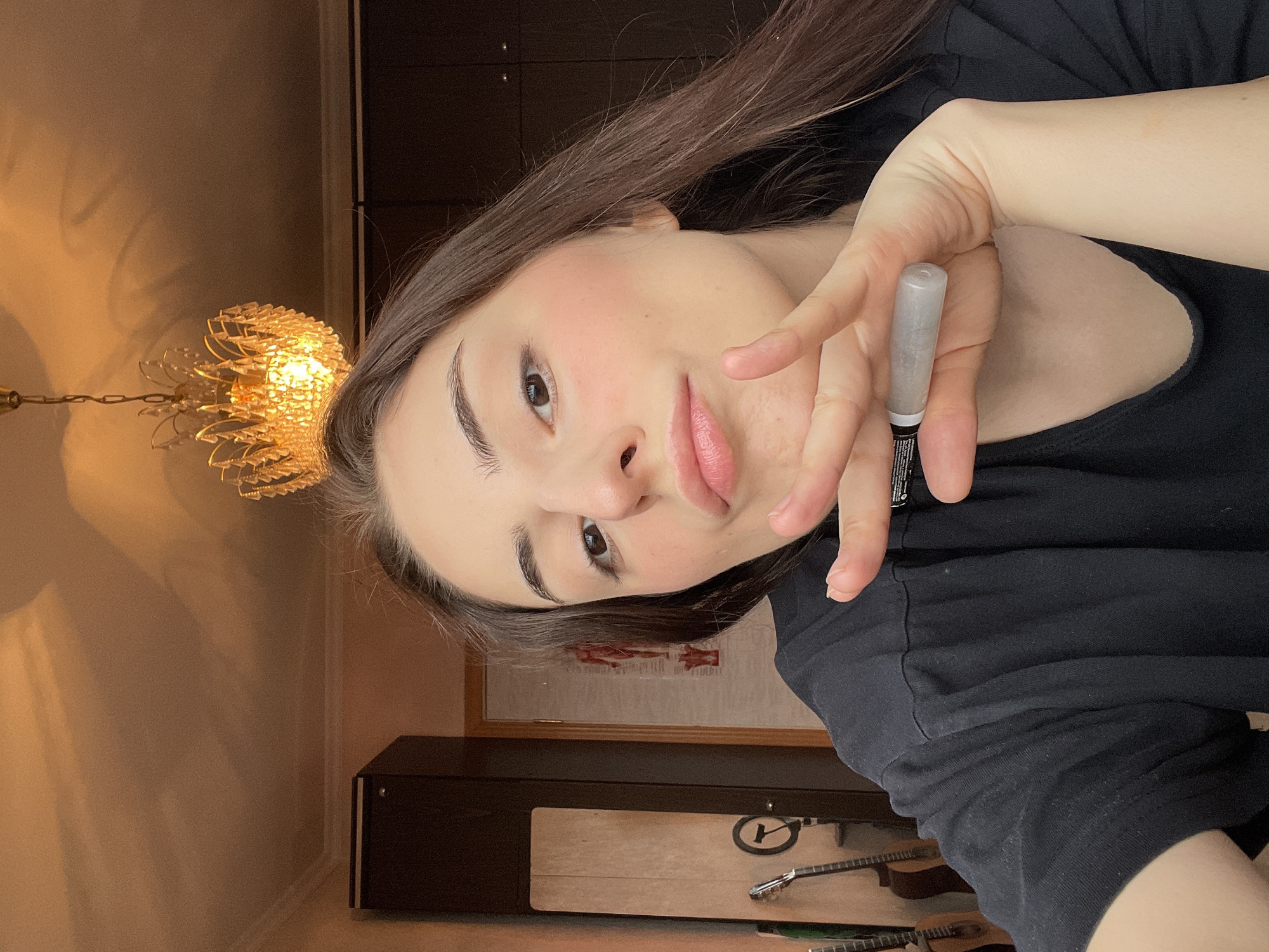 A young woman with long, dark hair and fair skin is holding a makeup applicator close to the camera. She is wearing a black t-shirt. The background includes a chandelier, a dark cabinet, and several guitars. The location cannot be definitively determined to be Moscow, Russia, based on the visual information provided.