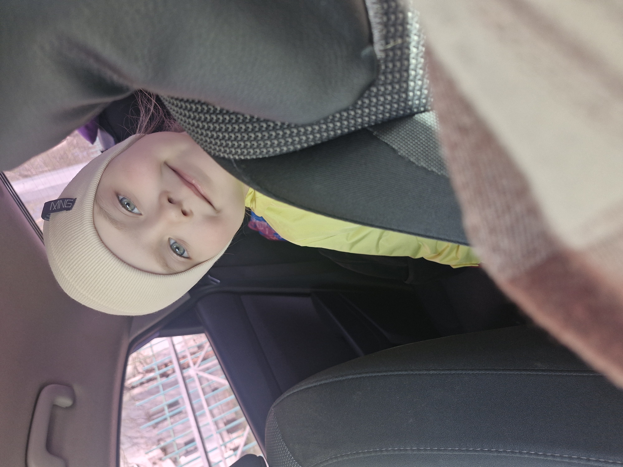 A child with light skin and blue eyes, wearing a cream-colored beanie with a visible tag, is positioned in what appears to be the backseat of a car. The child is smiling and appears to be looking up at the camera. The interior of the car is visible, featuring dark gray seats with stitching details. The window shows a blurred, indistinct view of a city building with a grid-like facade, suggesting an urban environment. A yellow and multi-colored garment is partially visible. There's also a draped blanket on the side. The overall impression is that the child is a passenger in a car in an unidentified city.