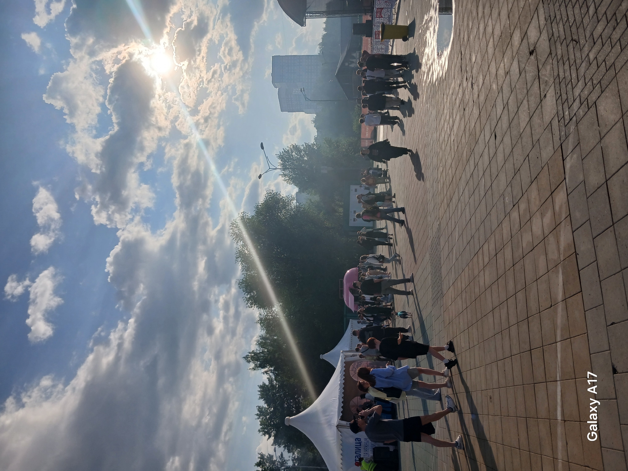 The image depicts a scene in Yekaterinburg, Russia. People are lined up outdoors on a brick-paved area. A white tent structure with writing on the side is visible on the left, possibly a booth or stall for an event. Further in the background are trees and what appears to be a building. The sky is partly cloudy with bright sunlight.