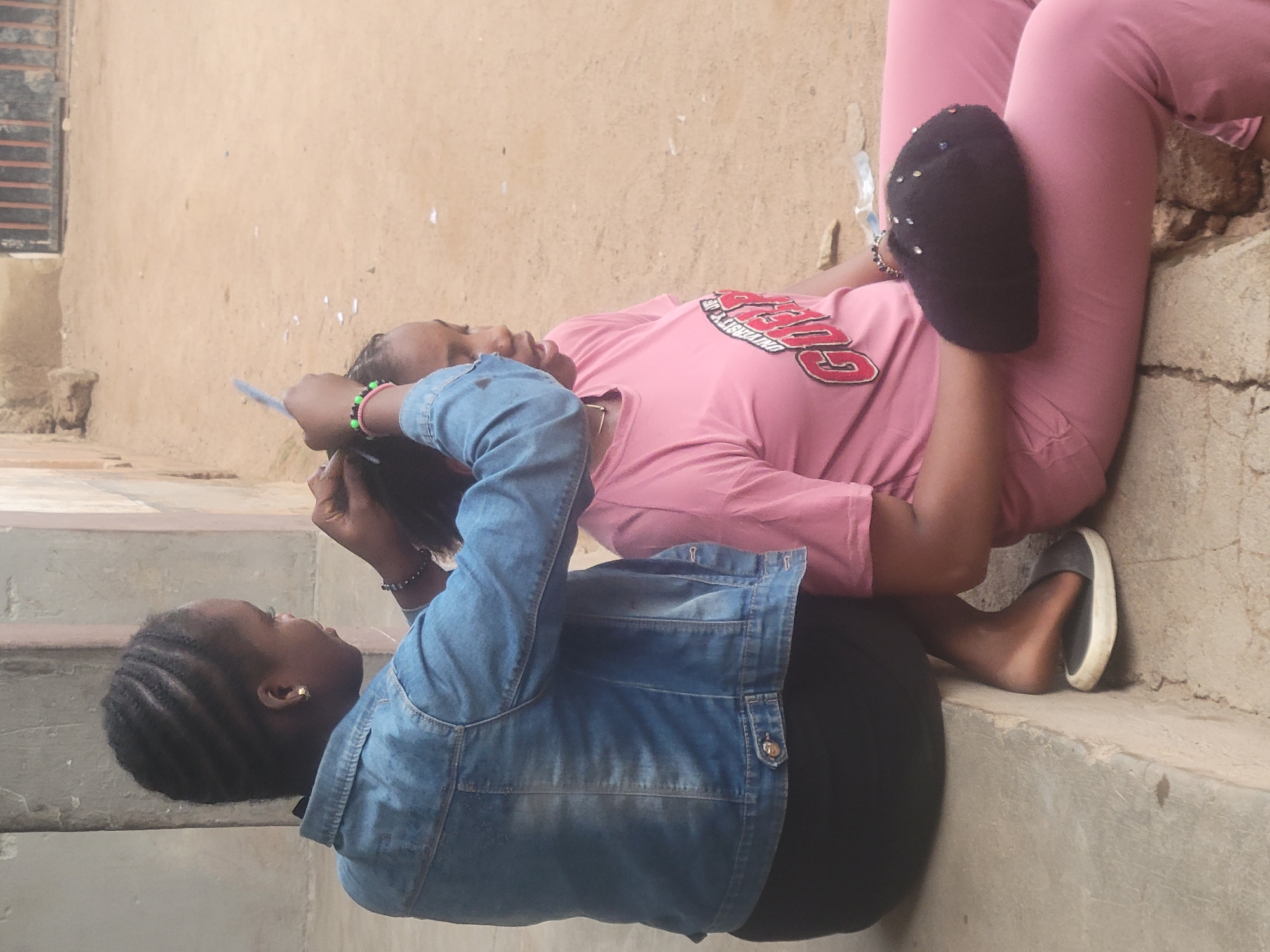 Two young women are sitting on a concrete step outdoors. One woman wears a light blue denim jacket and black pants. She is facing upwards and appears to be working on the hair of the other woman. The second woman is lying down with her head resting on the first woman's lap. She is wearing a pink t-shirt, pink pants, and one black slipper. The location is likely in Jos, Nigeria, based on the setting and the individuals present. The background shows a beige wall and a window with bars.
