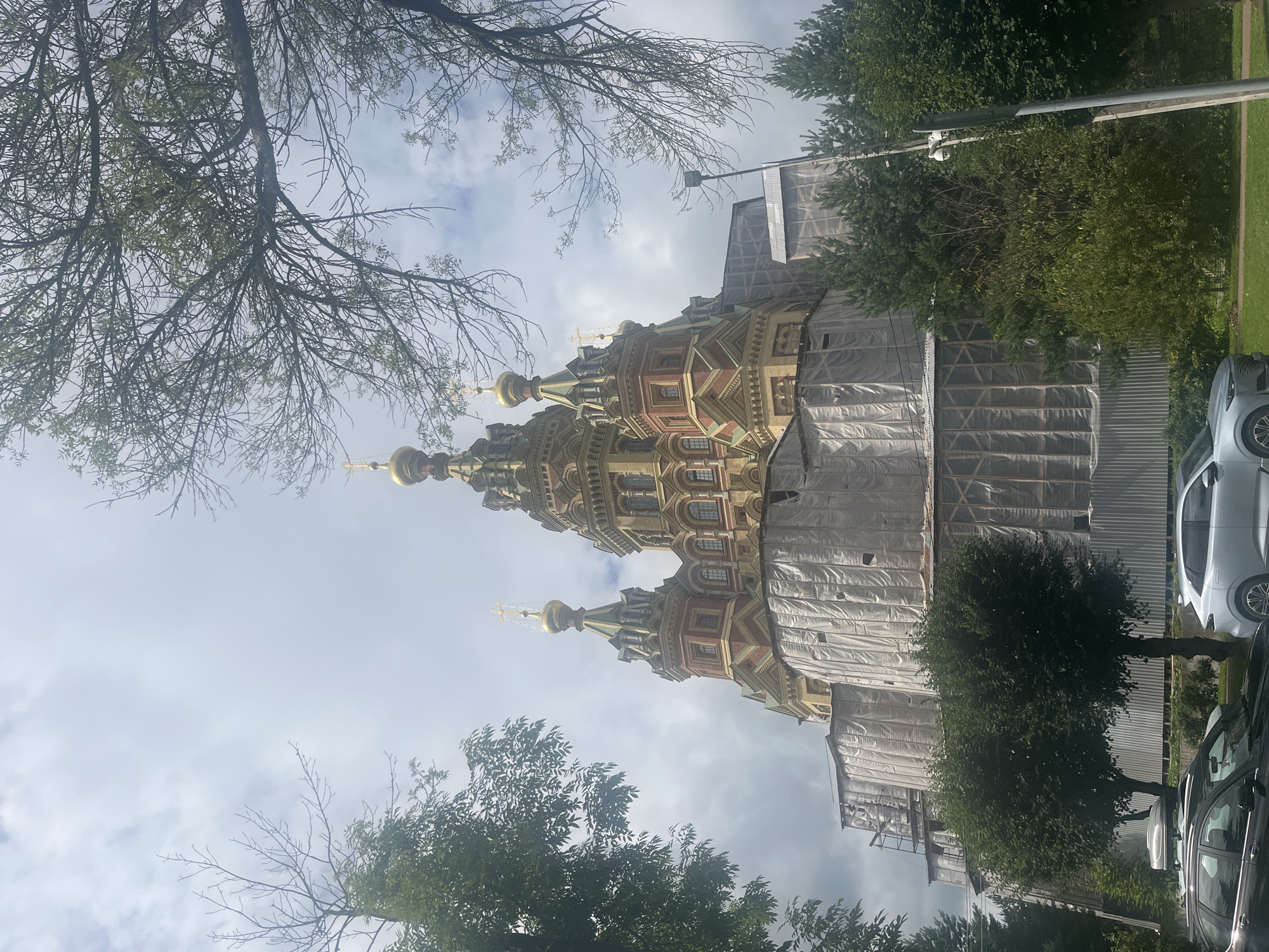 In Peterhof, Russia, the Petrine Baroque-style cathedral, likely the St. Peter and Paul Cathedral, is partially obscured by white tarpaulin and scaffolding, indicating ongoing renovation or preservation work. The cathedral features multiple golden onion domes topped with crosses. Lush green trees frame the structure, with some bare branches of a deciduous tree extending from the upper left corner. The sky is overcast with a mix of light and dark clouds. In the foreground, two cars are parked on a paved surface bordering a grassy area. A modern street lamp with a security camera is visible to the right of the cathedral. The scene suggests a daytime setting with overcast weather conditions.