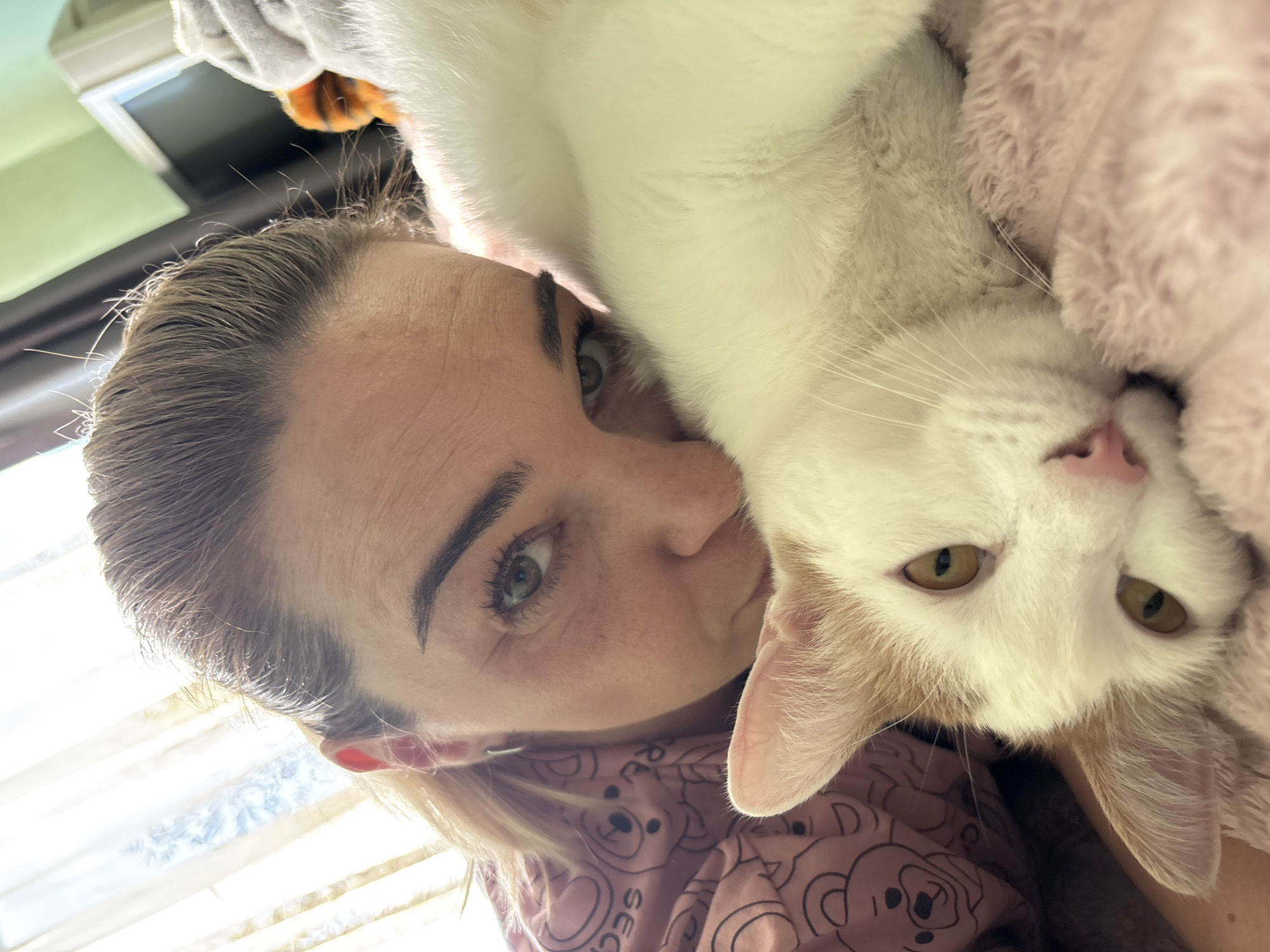 In a close-up, indoor, selfie-style shot, a woman with blonde hair tied back and wearing a pink pajama top with teddy bear prints leans her face towards a white and ginger cat. The woman's eyes are open and looking towards the camera, her brow furrowed slightly. The cat is lying down, nestled against the woman, with its head resting on a pink, fluffy surface. The cat's eyes are also open, and it appears relaxed. The background shows a window with sheer curtains and a glimpse of a television screen, suggesting the setting is likely a bedroom or living room in a residential area. The lighting is bright and natural, coming from the window. There are no discernible elements that can definitively place the scene in a specific city in Russia; the environment appears generic to a domestic setting.
