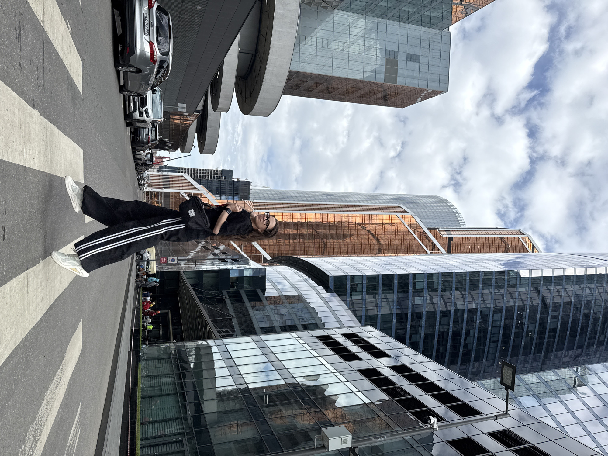 A woman stands on a crosswalk in Khimki, Russia. She wears sunglasses, black sweatpants with white stripes, and white sneakers. She is dressed in a black shirt and has a black bag slung over her shoulder. Buildings tower around her, reflecting the sky. Cars are parked along the street near the crosswalk.