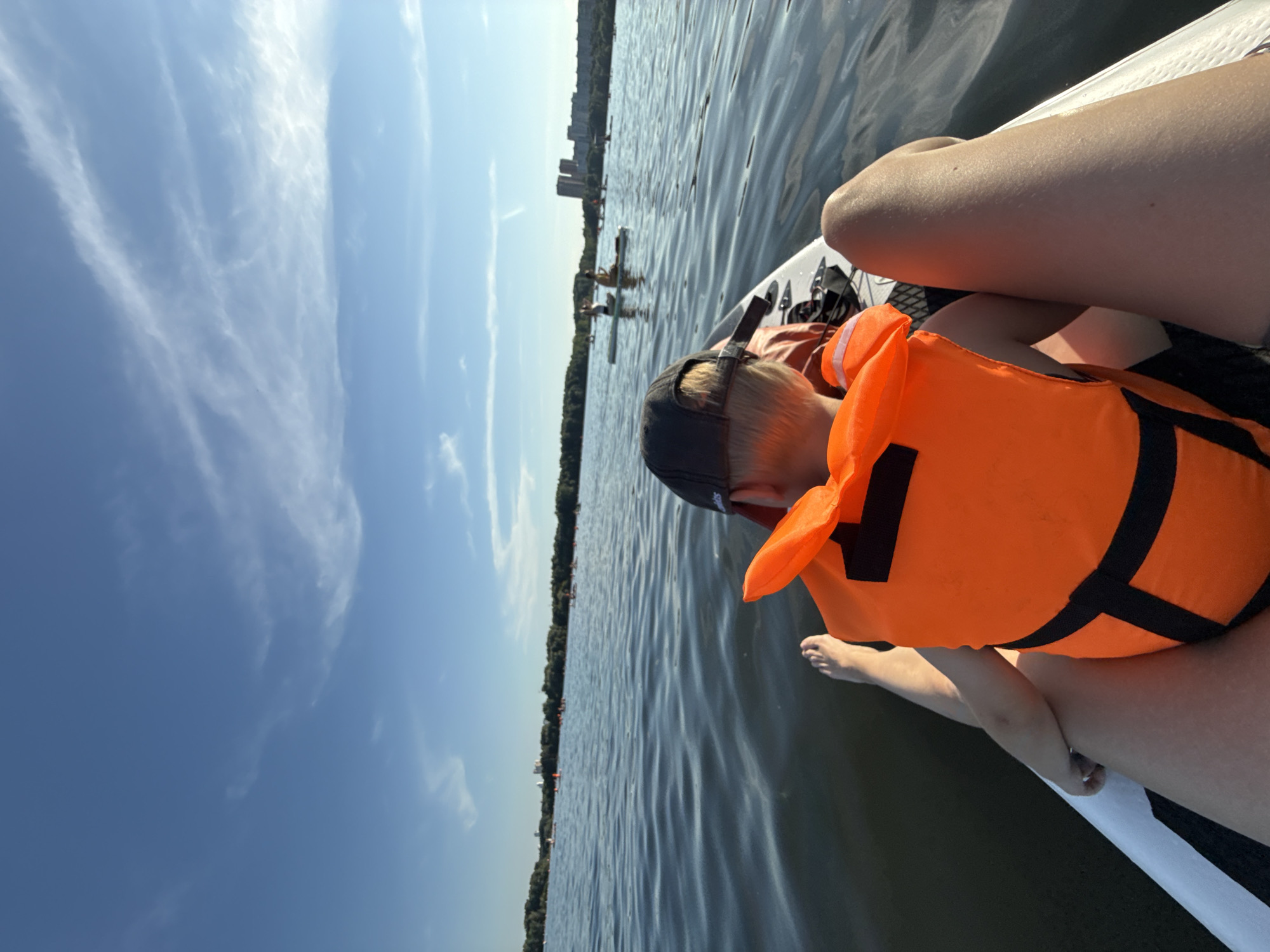 In this close-up, over-the-shoulder shot, a child wearing a bright orange life vest is seated on a paddleboard, facing away from the camera. The child's bare feet are visible, resting on the paddleboard. To the right, the legs of an adult are visible, also seated on the paddleboard. The paddleboard is white with black accents. The scene takes place on a body of water, likely a lake or large river, with gentle ripples on the surface. In the background, the tree-lined shore of a city is visible under a bright blue sky with scattered white clouds. Several other paddleboards with people on them are scattered across the water in the mid-ground. The overall impression is a recreational outing on a sunny day.