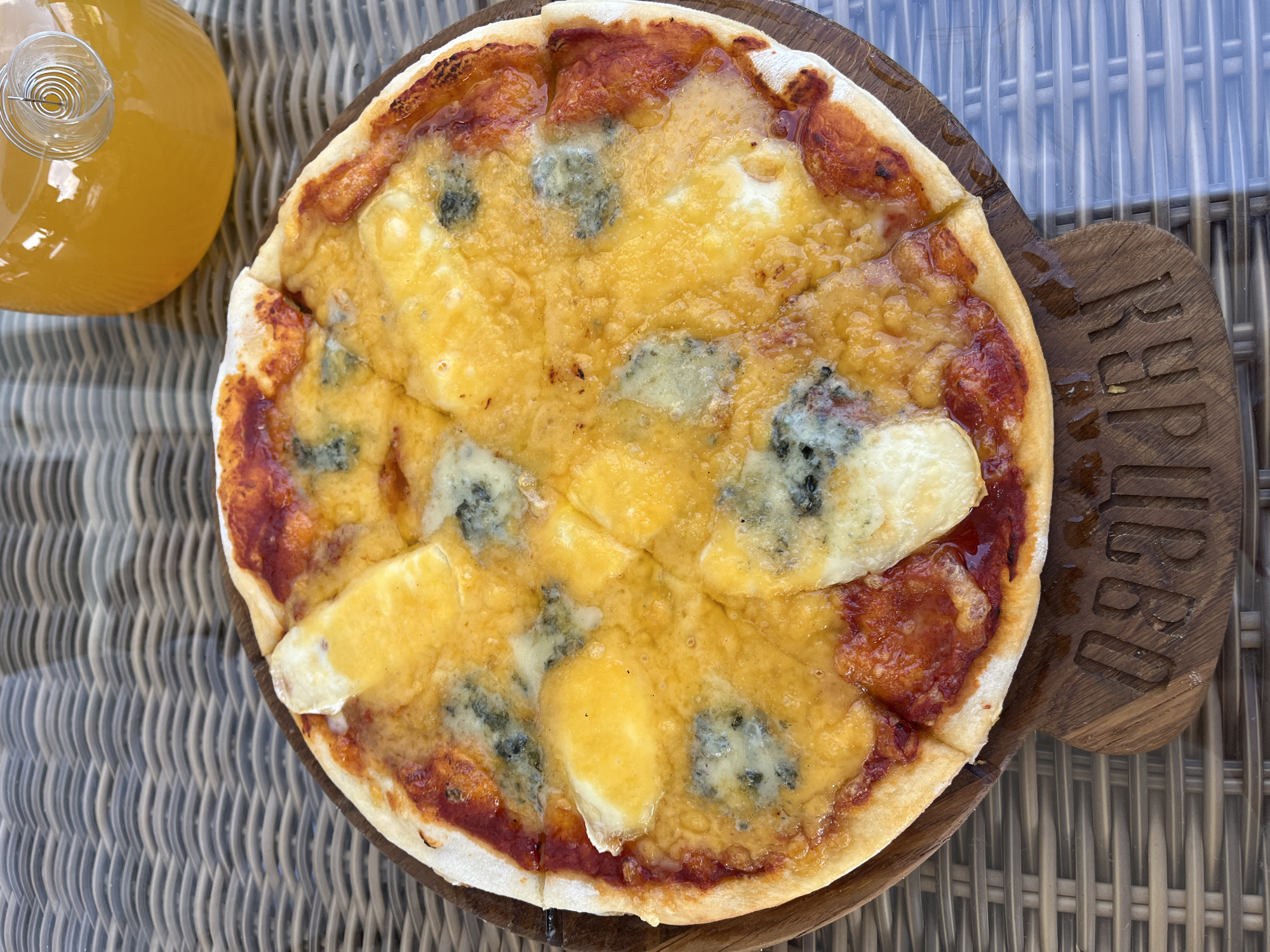An overhead shot captures a small, round pizza on a wooden board with a carved inscription. The pizza is topped with melted cheese, including a yellow variety and pieces of blue cheese, all on a bed of red sauce. The crust is lightly browned. To the upper right of the pizza is a clear glass bottle filled with an orange beverage, possibly juice, with a silver coil-like object inside. The pizza and bottle rest on a woven, wicker-like surface, likely a table. The inscription on the wooden board appears to be "KRPUGOV". The scene is set outdoors, suggested by the diffused natural lighting. No people are visible in the image.