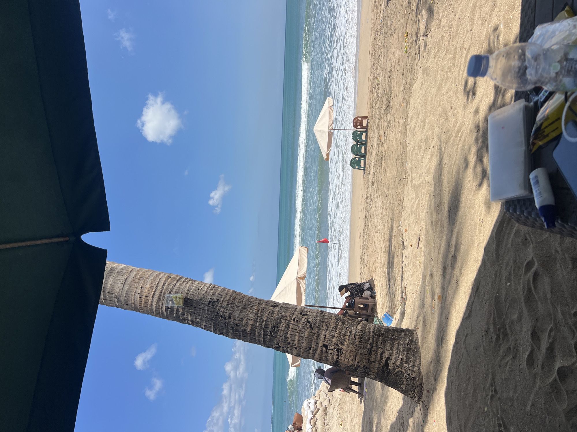 A tropical beach scene in Kuta, Indonesia, unfolds under a clear blue sky dotted with white clouds. A large palm tree dominates the foreground, its textured trunk casting a shadow on the sandy ground. In the middle ground, two beach umbrellas provide shade, with a few empty plastic chairs arranged beneath one of them. Further out, the ocean stretches to the horizon, with gentle waves breaking on the shore. In the background, a red flag is planted in the sand, and a few people are visible sitting or standing on the beach, some beneath umbrellas. The foreground also features a table with a plastic water bottle, a packet of tissues, and a wallet. The overall impression is one of a relaxed beach atmosphere, likely a popular spot for tourists and locals alike.