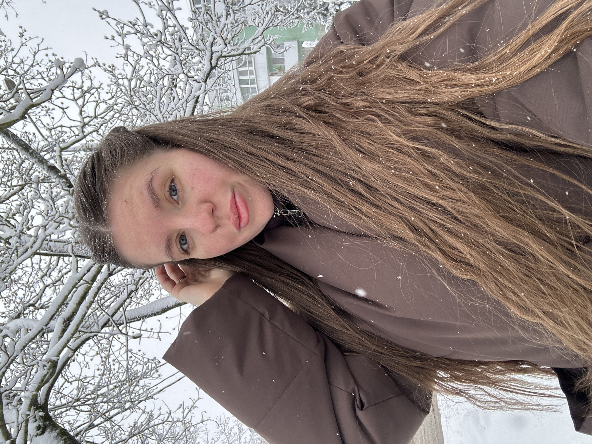 A medium-close-up, diagonally oriented shot captures a young person, likely female, outdoors in a snowy environment. The subject has long, flowing light brown hair, which is visibly dusted with numerous small white snowflakes. She wears a dark brown, possibly padded, winter jacket, and looks directly at the camera with a soft, slightly smiling expression. Her fair skin shows a hint of pink on the cheeks, and her eyes are blue. Her left hand is subtly resting near her ear or cheek.
The background is dominated by a dense network of snow-covered tree branches, suggesting a winter landscape. Through the bare, snow-laden branches, a light-colored building, possibly an apartment block, with visible windows can be seen. Small, distinct snowflakes are suspended in the air, indicating ongoing snowfall. The overall lighting suggests an overcast winter day, consistent with a snowy scene in Klin, Russia. No visible text is present in the image.