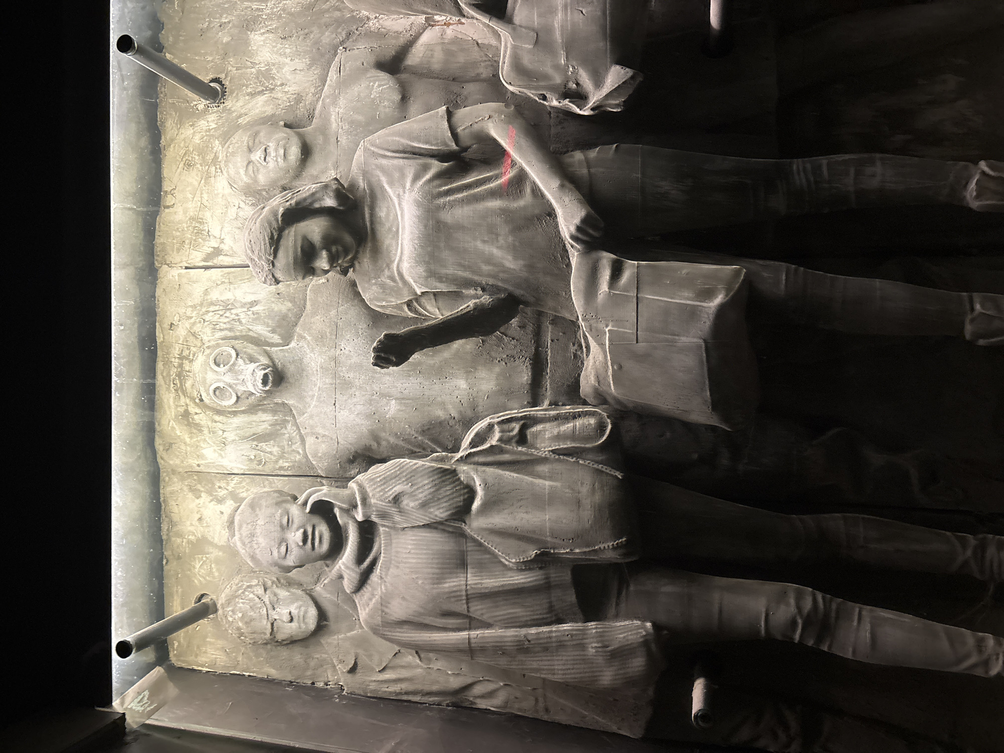 The image shows a bas-relief sculpture depicting figures in distress, likely related to a historical event. The sculpture appears to be made of a light-colored material, possibly concrete or plaster. Several figures are depicted in detail. At least one figure is wearing a gas mask. There are two metal poles attached to the edge of the sculpture. The image was taken at the Memorial Church "St. Alexander Nevsky" in Sofia, Bulgaria.