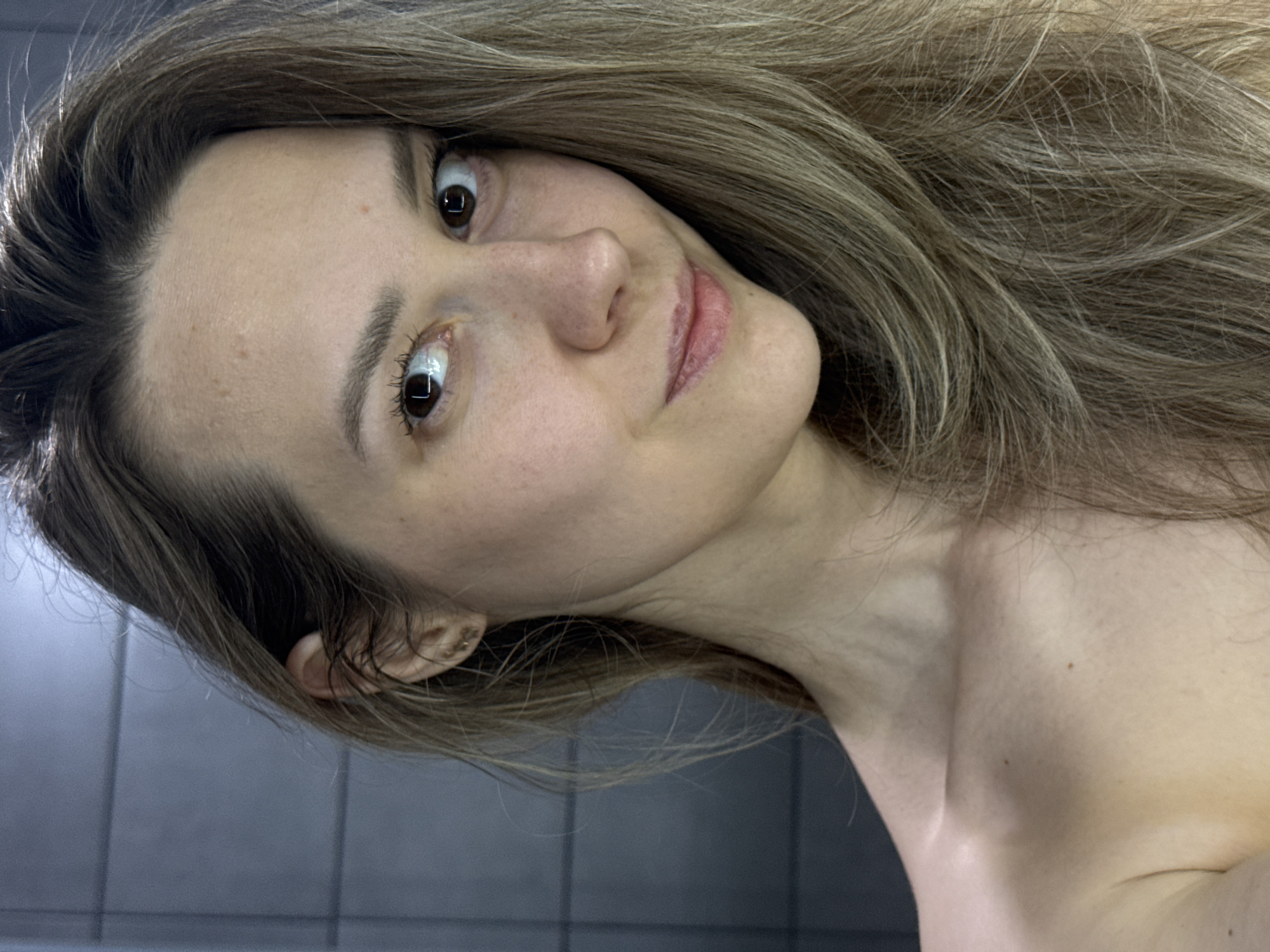 A fair-skinned woman with shoulder-length, light brown hair is centered in the image, positioned at a tilted angle. Her facial features include brown eyes, a straight nose, and slightly parted lips. She appears to be looking directly at the camera with a slight smile. She is wearing earrings in her left ear. The background consists of what appears to be a tiled wall, with dark gray horizontal tiles. There is no indication of this image being taken in Chaikovsky, Russia.