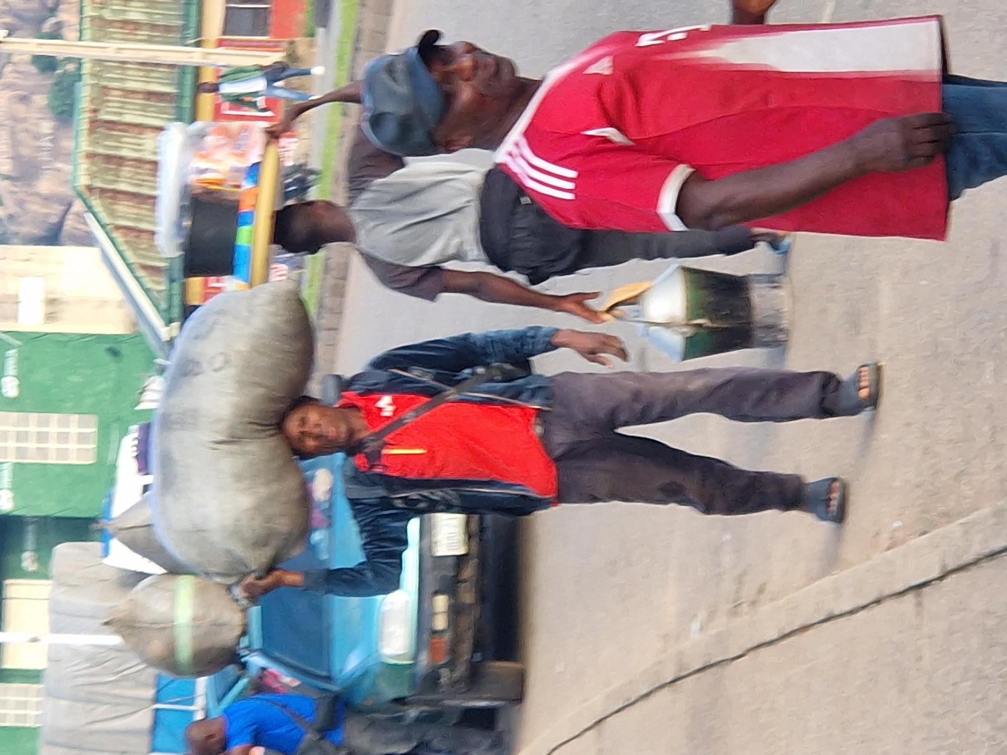A man in the foreground is wearing a red shirt with white stripes on the sleeves and a black baseball cap. He is looking to his right. Behind him, another man is carrying a large sack on his head, wearing a dark jacket over a red shirt and dark pants. A teal-colored car is parked behind him. In the background, there are buildings with corrugated metal roofs and a distant hill. Other individuals are visible in the background, and the scene appears to be on a street in an urban environment.