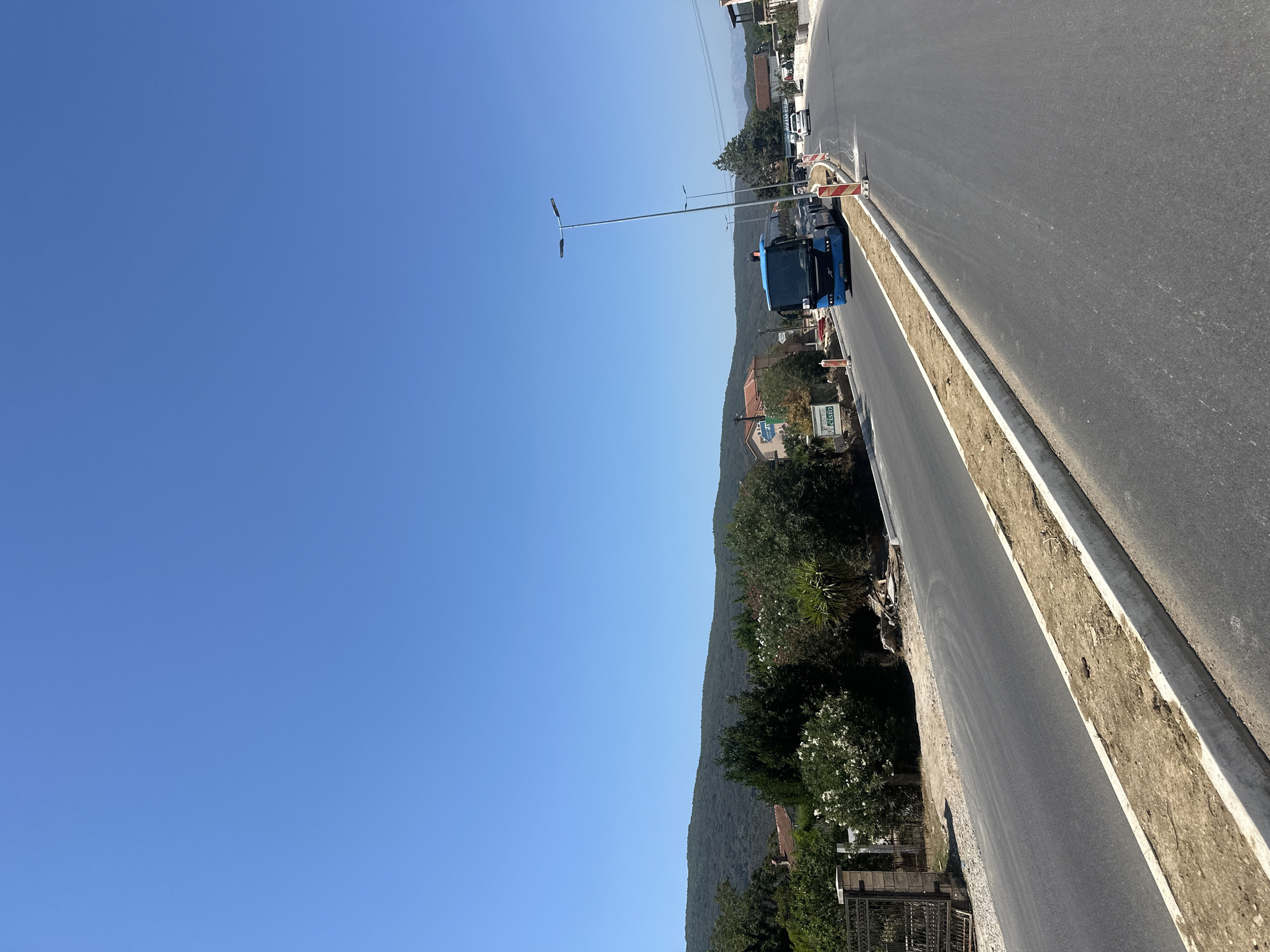 The image depicts a clear, bright day in Lastva Grbaljska, Montenegro, with a vast expanse of blue sky dominating the upper portion. In the mid-ground, a blue bus is visible traveling on a paved road. The road appears to be undergoing construction or refurbishment, with concrete curbs and loose dirt visible along its edge. Several streetlights are positioned along the roadside, one of which is notably tall. In the background, a verdant, hilly landscape is present, with scattered buildings, some featuring red-tiled roofs, nestled amongst trees and vegetation. The overall scene suggests a rural or semi-rural setting within the city, with ongoing development evident.